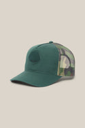 The 686 Mid-Pro Wreath Unstructured 5 Panel Mesh Hat by 686 is a green polyester cotton woven men's trucker hat, featuring a solid green front panel with a black embroidered logo, a matching green bill, and camouflage mesh back panels. It is shown against a plain beige background.