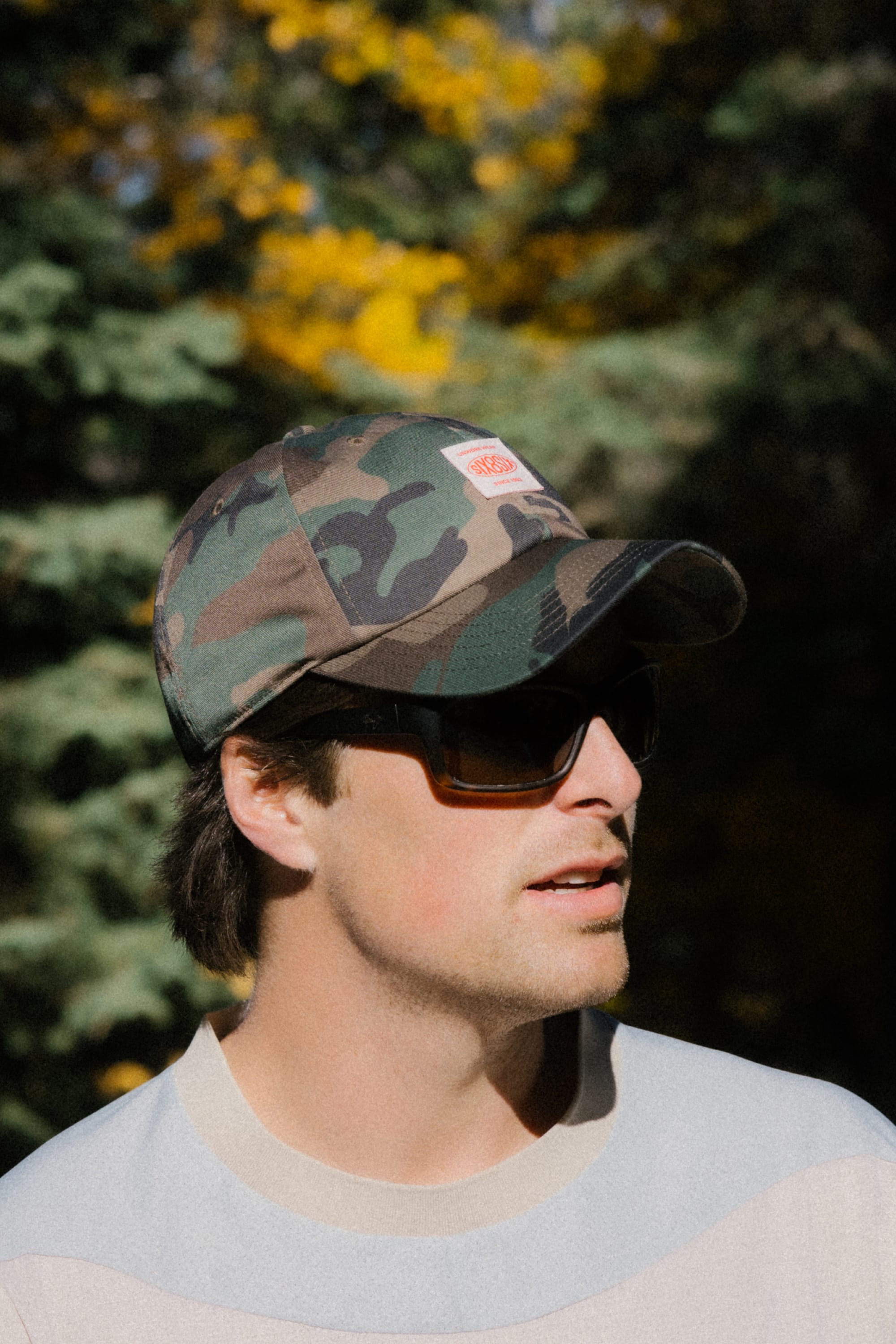 A person wearing the 686 Low-Pro Unwork Unstructured 6 Panel Hat by 686—a men's camouflage baseball cap—and dark sunglasses stands outdoors, with green trees and yellow foliage blurred in the background.