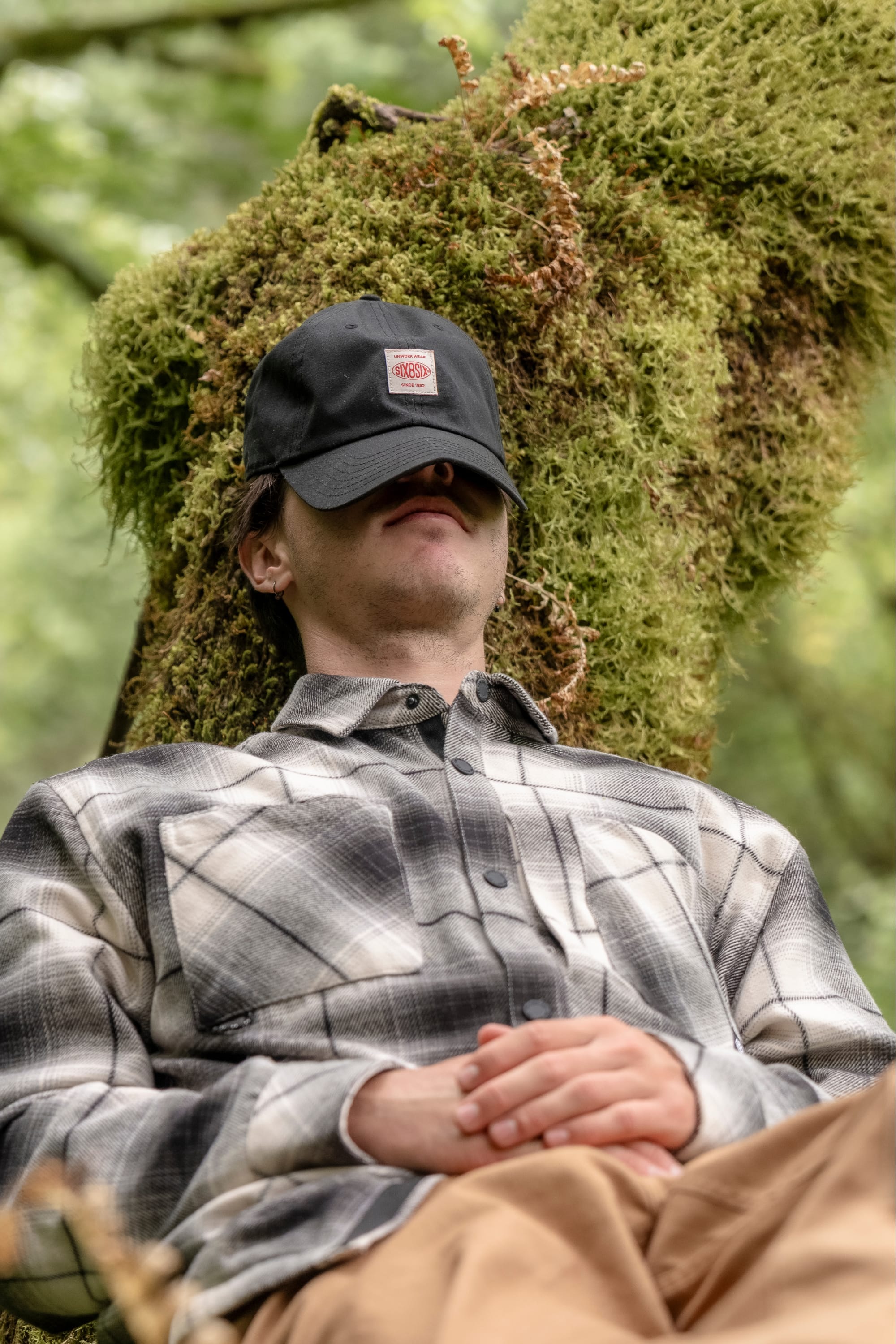 A person wearing a plaid shirt and the 686 Low-Pro Unwork Unstructured 6 Panel Hat by 686 sits against a mossy tree, eyes shaded by the hat's brim and hands folded, appearing relaxed in a forest setting.