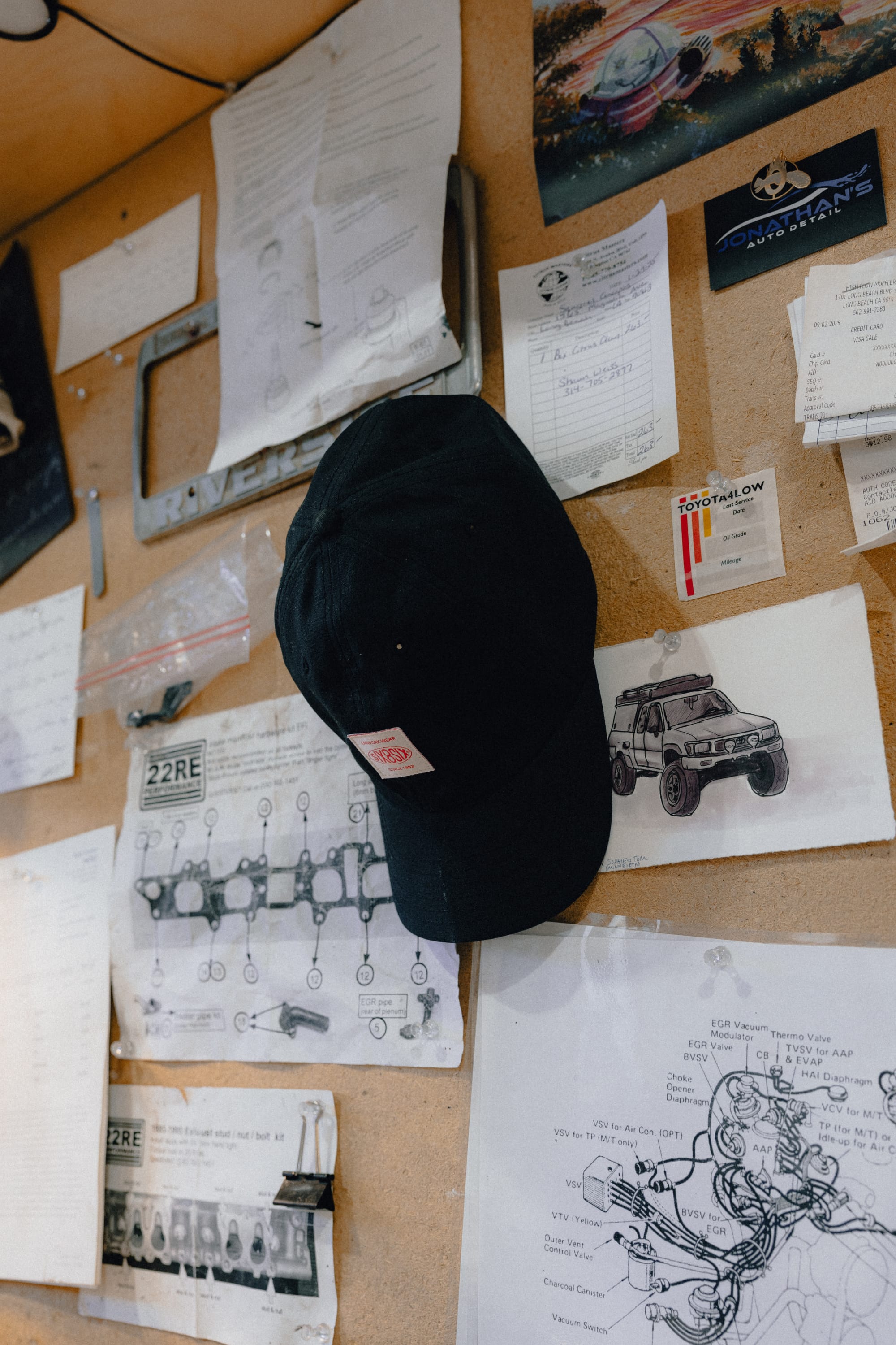 A 686 Low-Pro Unwork Unstructured 6 Panel Hat in black by 686 hangs on a corkboard covered with automotive diagrams, handwritten notes, car sketches, receipts, and business cards.
