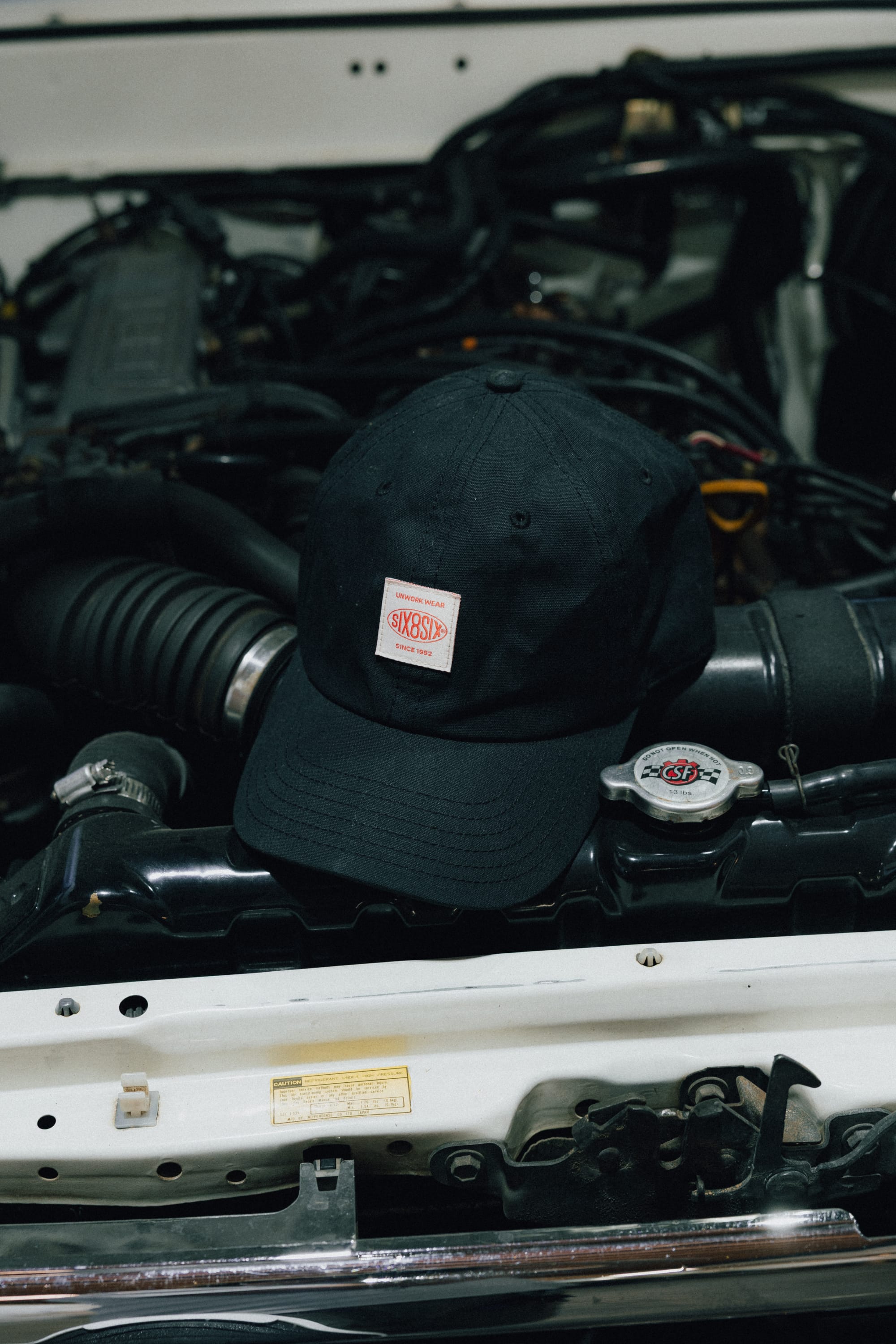 A 686 Low-Pro Unwork Unstructured 6 Panel Hat by 686, featuring a black polyester-cotton design and a white and red patch, rests on the open engine of a car, with various engine components and the radiator cap visible in the background.