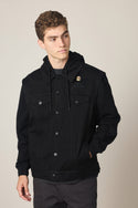 A young man with short curly hair wears the 686 Men's Metalhead Trucker Jacket by 686. The black woven jacket has a hood and a pin on the left chest. He has his left hand in his pocket and looks slightly to the left with a neutral expression.