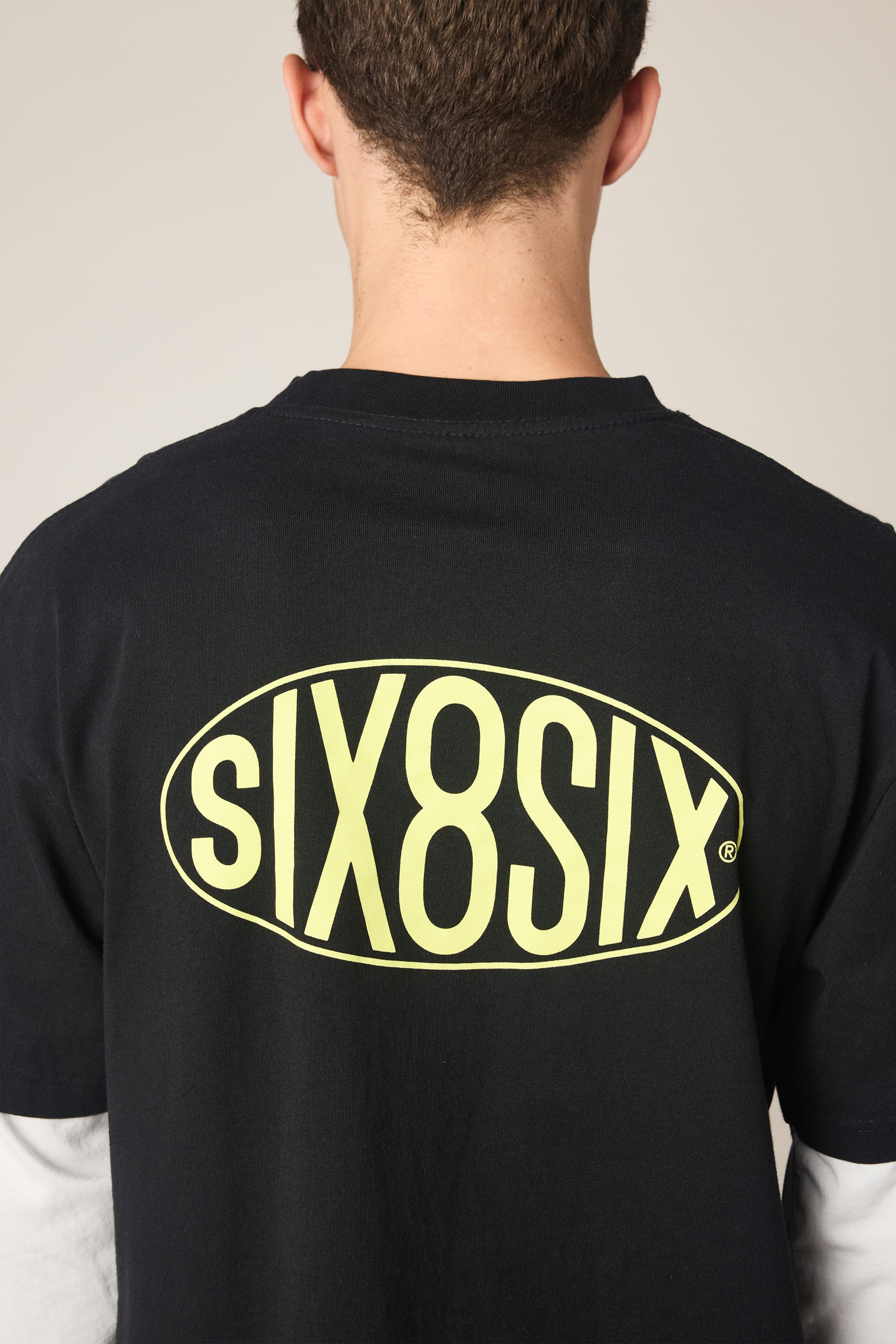 A person is seen from behind wearing the 686 Men's 92 Relaxed LS Double Trouble Tee in black, featuring bold yellow SIX8SIX lettering on the back and white sleeve layers for a stylish, relaxed look. Brand: 686.