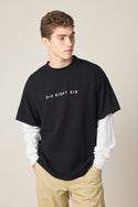 A young man with curly hair wears the 686 Wordmark Relaxed 2fer LS Tee by 686 over beige pants, glancing to the side with his hands in his pockets against a plain background.