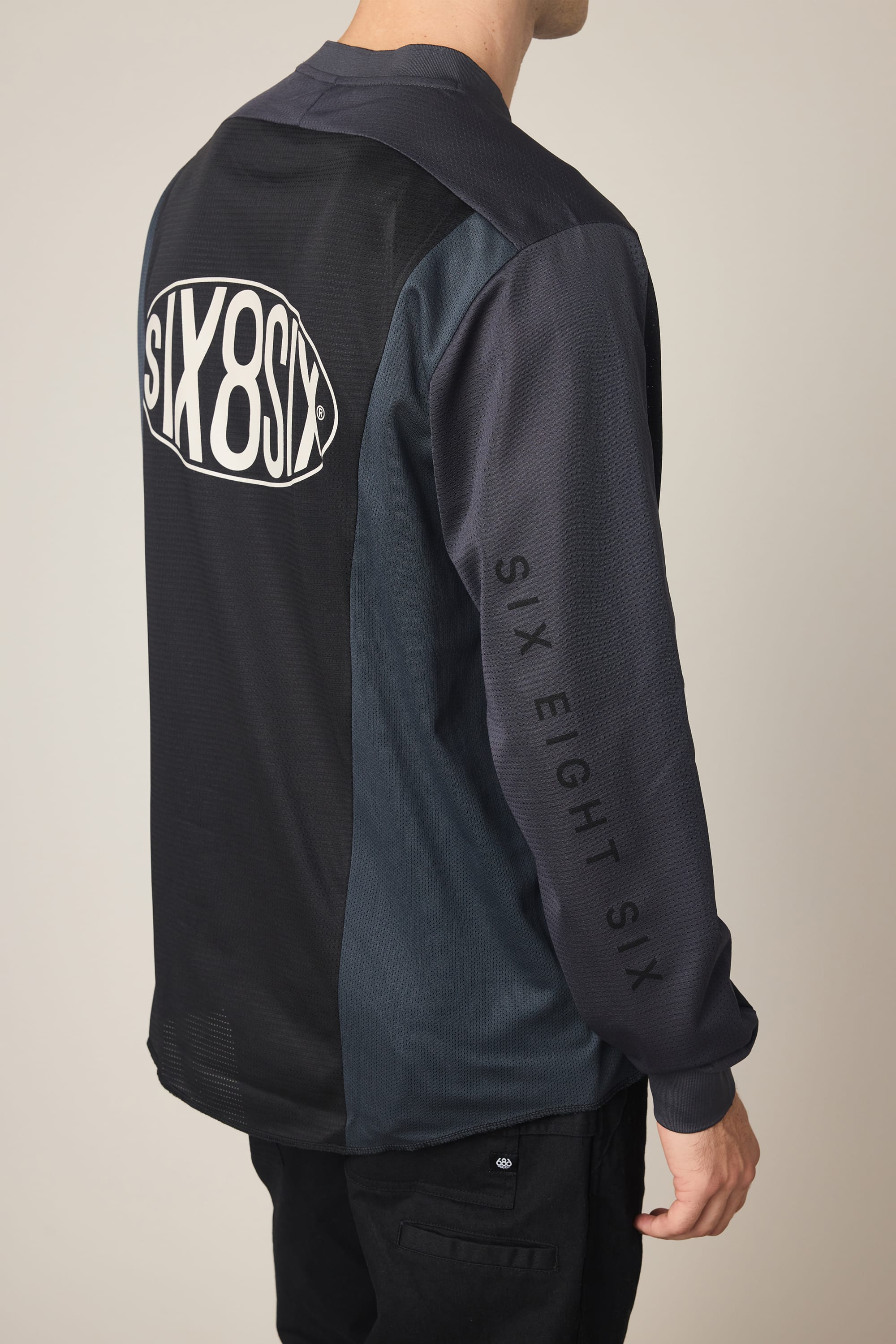 A person wearing the 686 Men's 92 Turbo LS Tech Tee, a dark long-sleeve polyester t-shirt by 686 featuring "SIX8SIX" on the back and "SIX EIGHT SIX" on the sleeve, stands with their back to the camera.