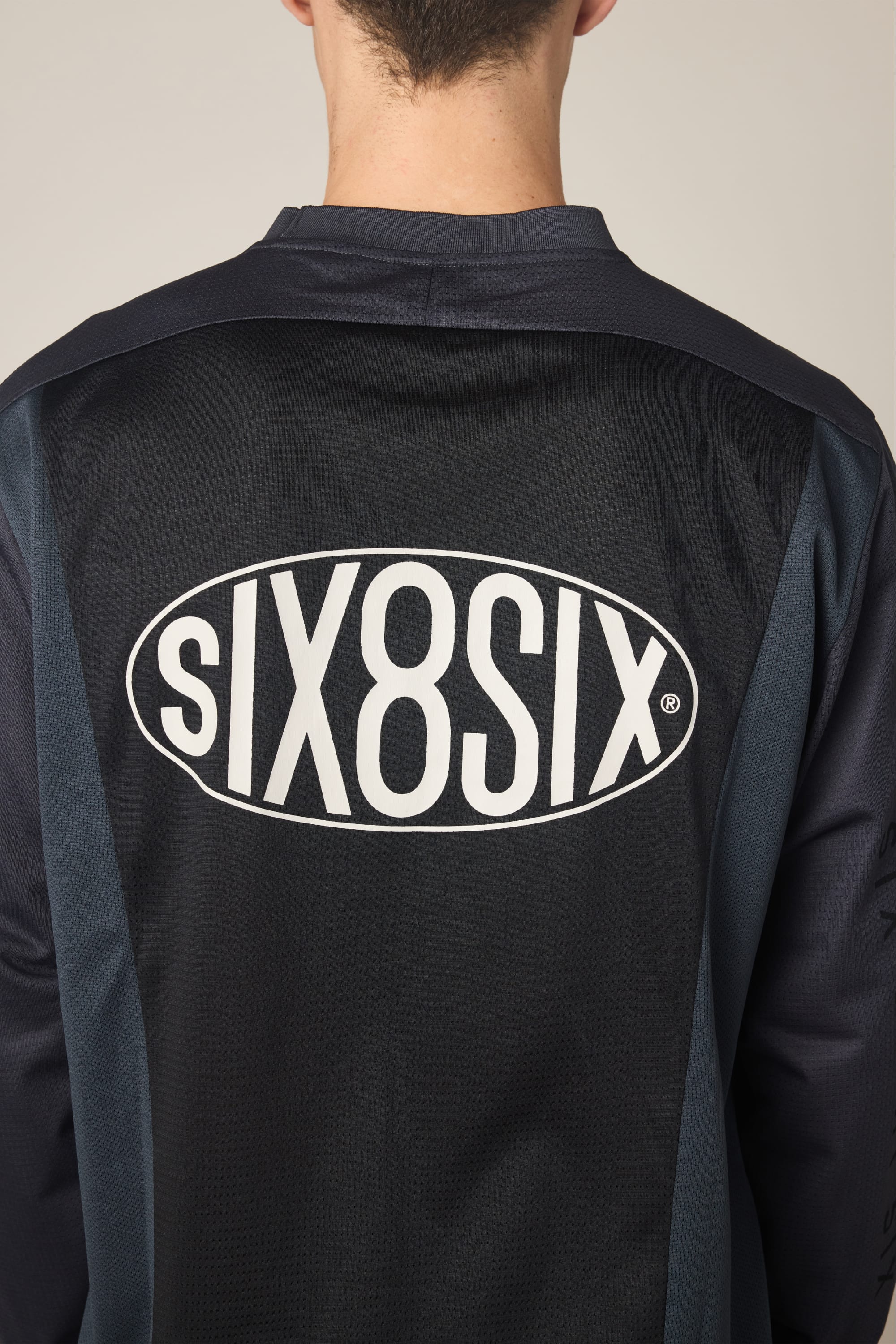 Person wearing a 686 Men's 92 Turbo LS Tech Tee in black and dark gray polyester, featuring a large white “686” logo inside an oval on the back. The focus is on the upper back and shoulders.