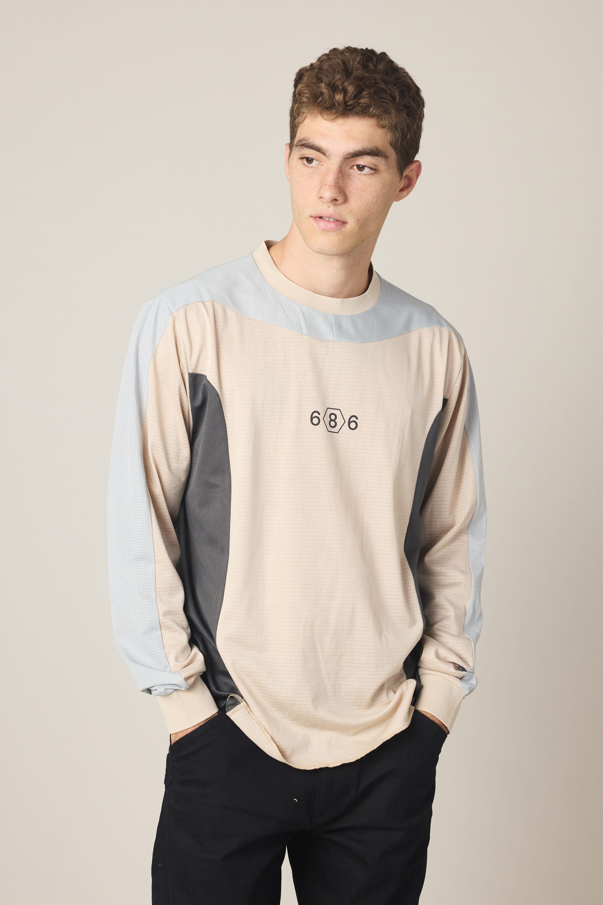 A young man with short curly hair wears the 686 Men's Hex Turbo LS Tech Tee by 686 in beige, gray, and blue, paired with black pants. He stands against a plain light background, hands in his pockets, looking to the side.