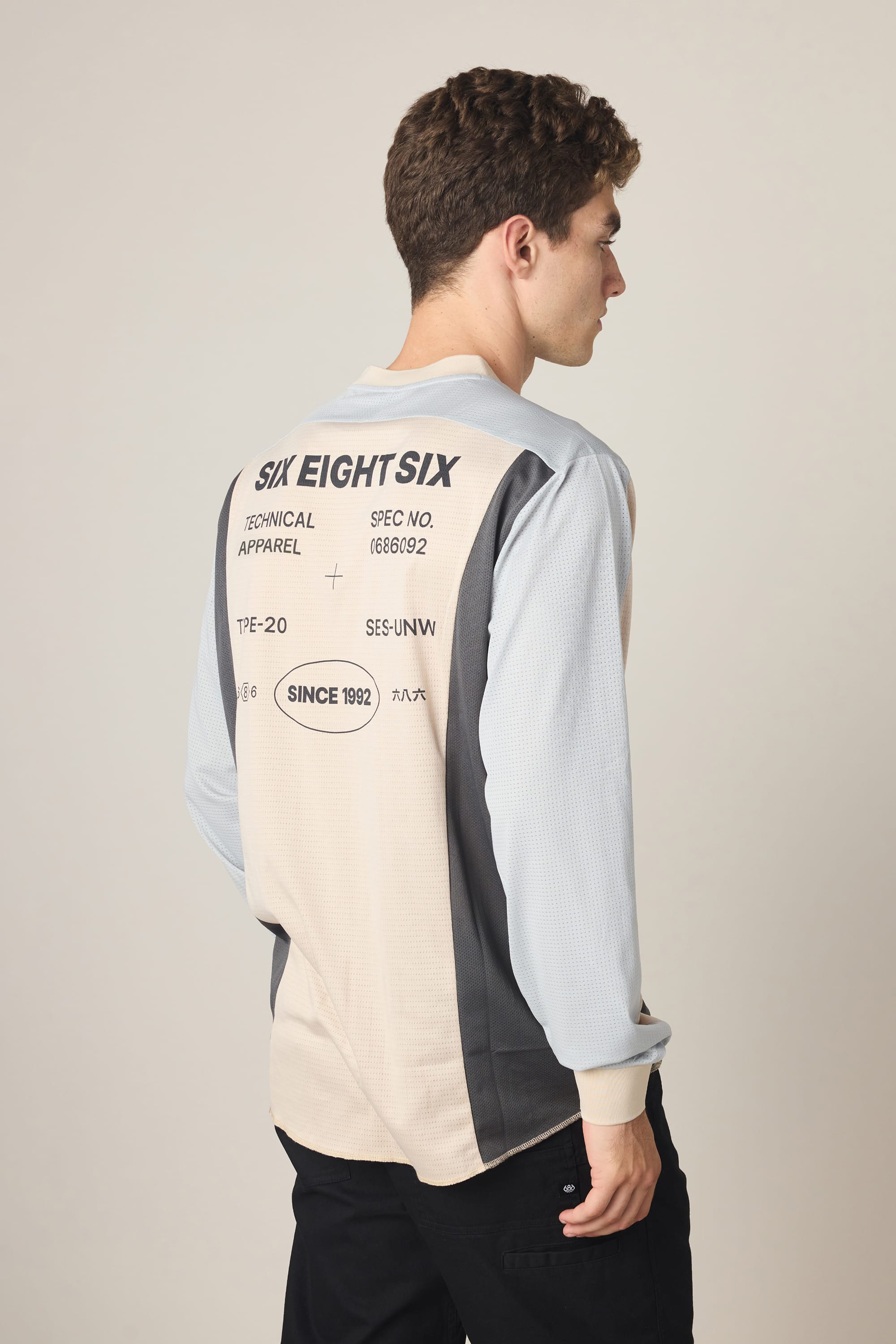 A young man with short curly hair stands facing away, wearing the 686 Men's Hex Turbo LS Tech Tee by 686. The polyester shirt features color-blocking and printed text like "SIX EIGHT SIX" and "SINCE 1992" on the back.