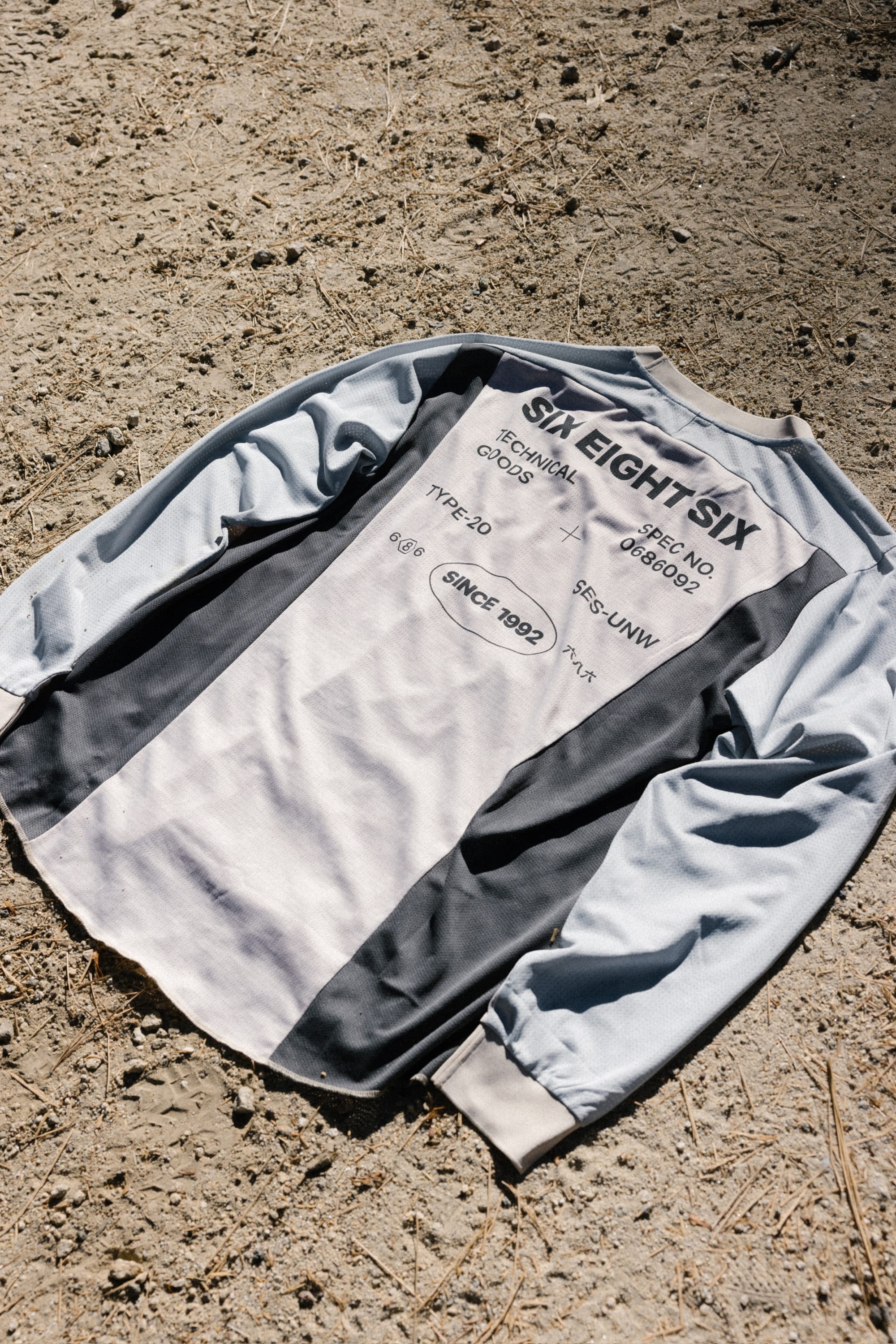 The 686 Men's Hex Turbo LS Tech Tee, featuring black and white panels, lies on dry, rocky ground. This long-sleeve t-shirt from 686 displays printed text such as SIX EIGHT SIX, technical details, and SINCE 1992. Made from 100% polyester for comfort and durability.