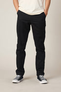 A person stands with hands in their pockets against a plain light background, wearing a white T-shirt, 686 Men's Everywhere® Standard Pant - Slim Fit by 686, and black-and-white sneakers. Their head is not visible in the image.