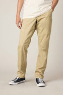 A person wears 686 Men's Everywhere® Standard Pant - Slim Fit in beige, a white T-shirt, and black low-top sneakers, standing with one hand in their pocket against a plain light background. The face is not visible.