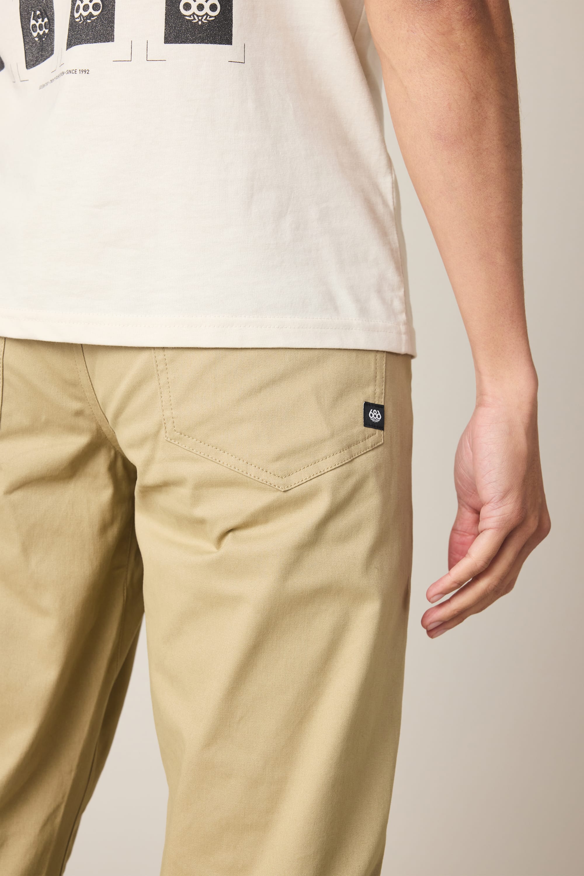 A person stands with their arm relaxed, photographed from behind and slightly to the side, wearing a white t-shirt and 686 Men's Everywhere® Standard Pant - Slim Fit in beige, featuring a black logo tag on the back pocket.