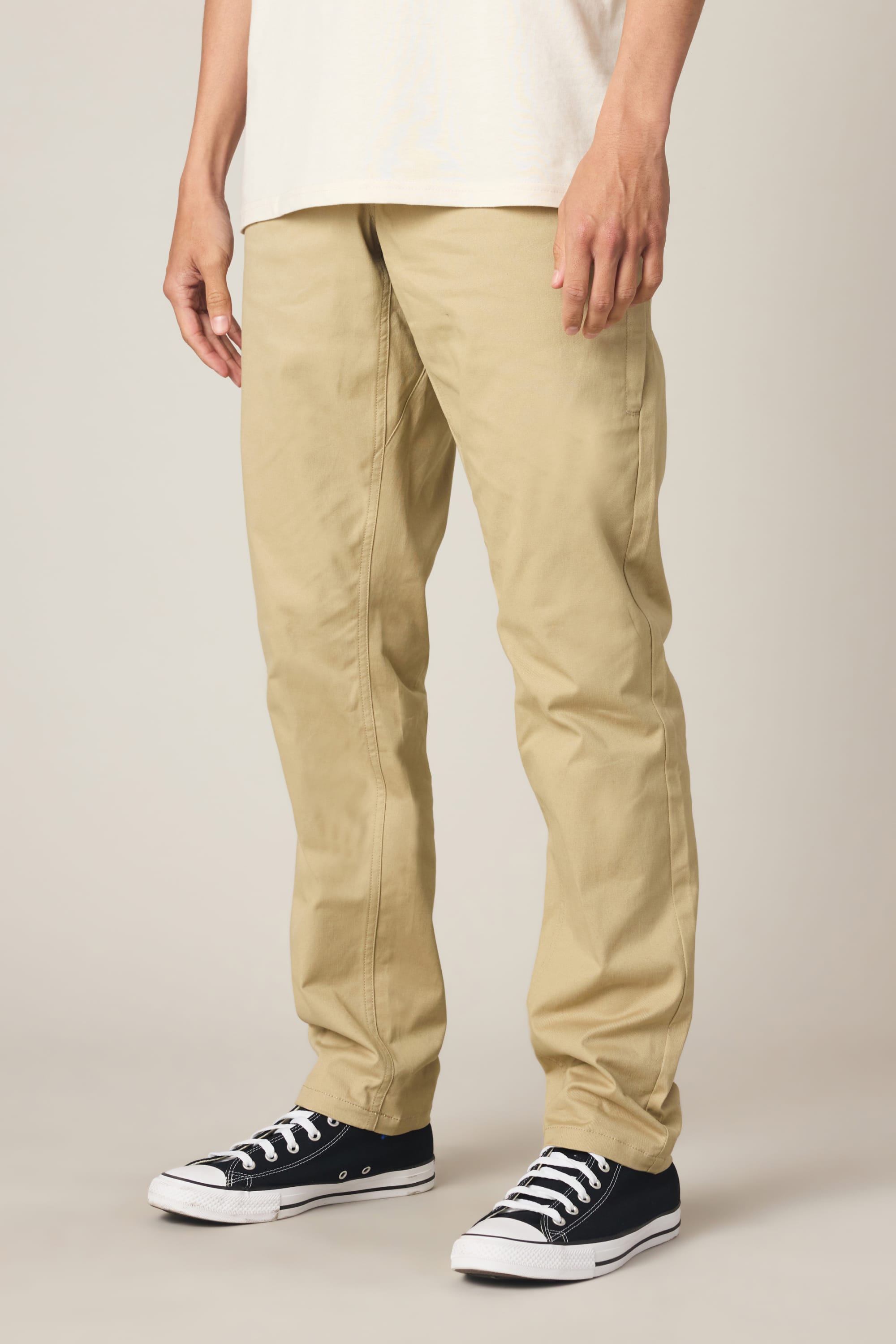 A person wearing 686 Men's Everywhere® Standard Pant - Slim Fit in light khaki, a white shirt, and black low-top sneakers stands against a plain background. Only the lower torso, legs, and feet are visible.