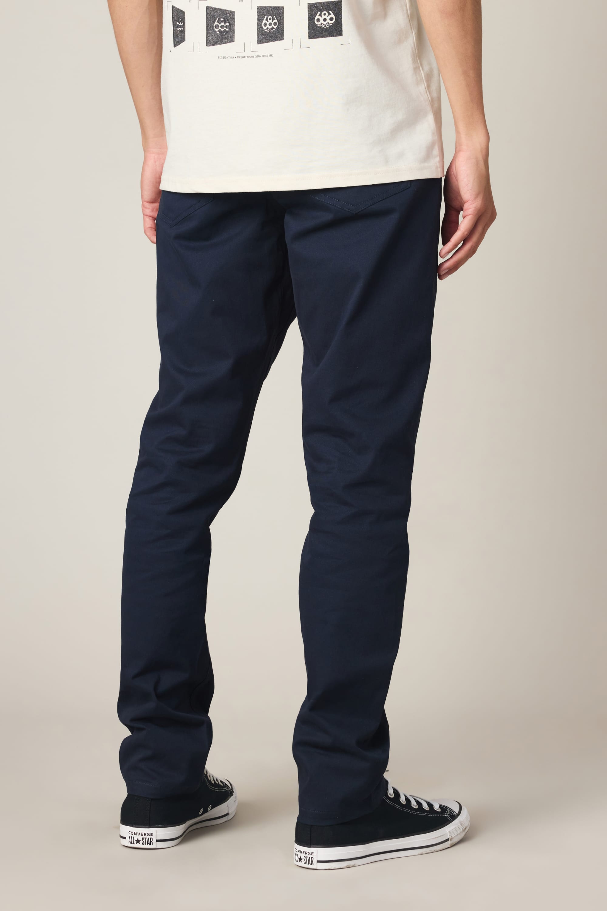A person stands facing away, wearing a white T-shirt, the 686 Men's Everywhere® Standard Pant - Slim Fit in navy blue by 686, and black low-top sneakers with white soles, against a plain light background.
