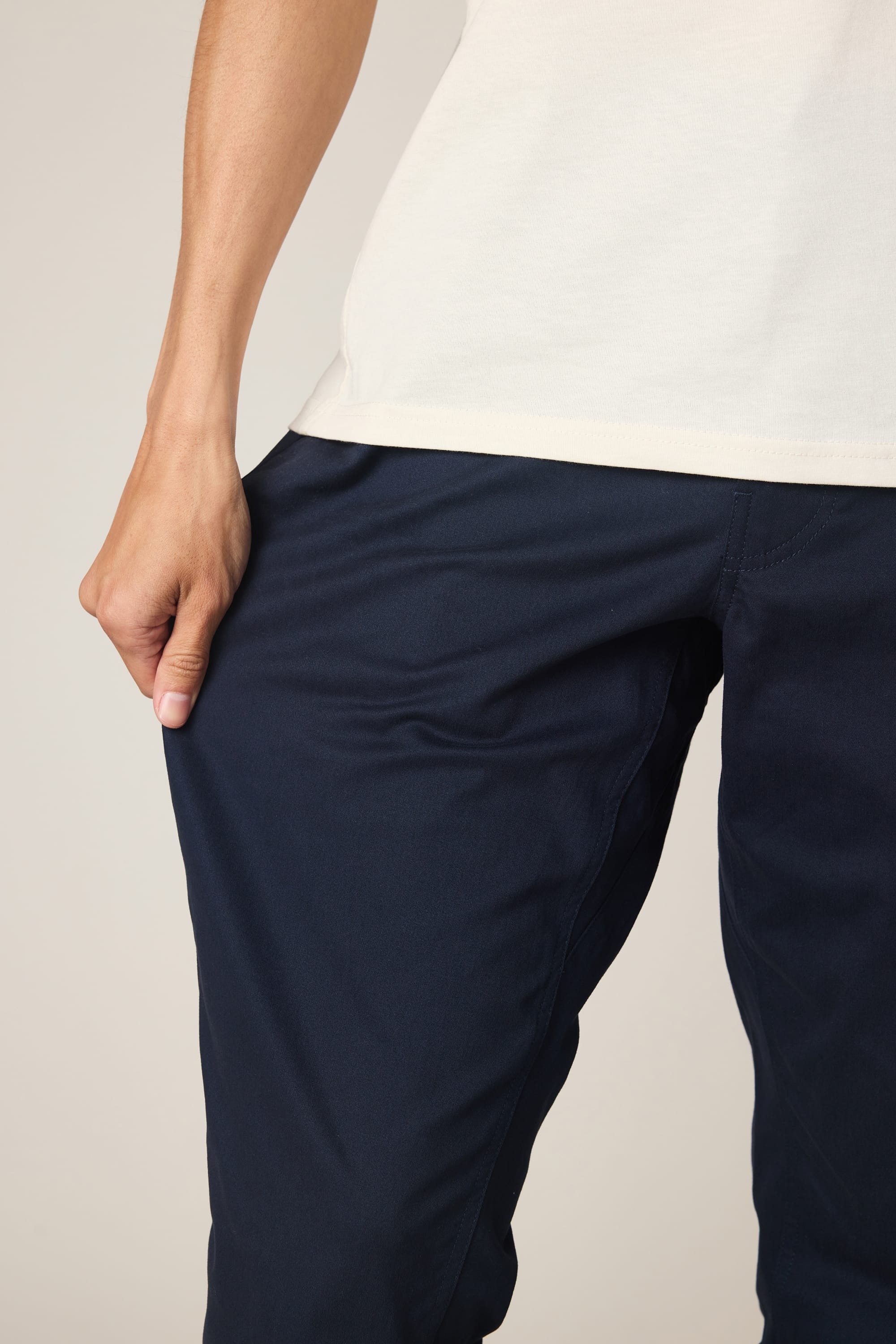 Wearing a cream shirt and 686 Men's Everywhere® Standard Pant - Slim Fit in navy, the individual stands with one hand on their hip, gently pulling the fabric of the pants outward. The image is cropped from the shoulders down.