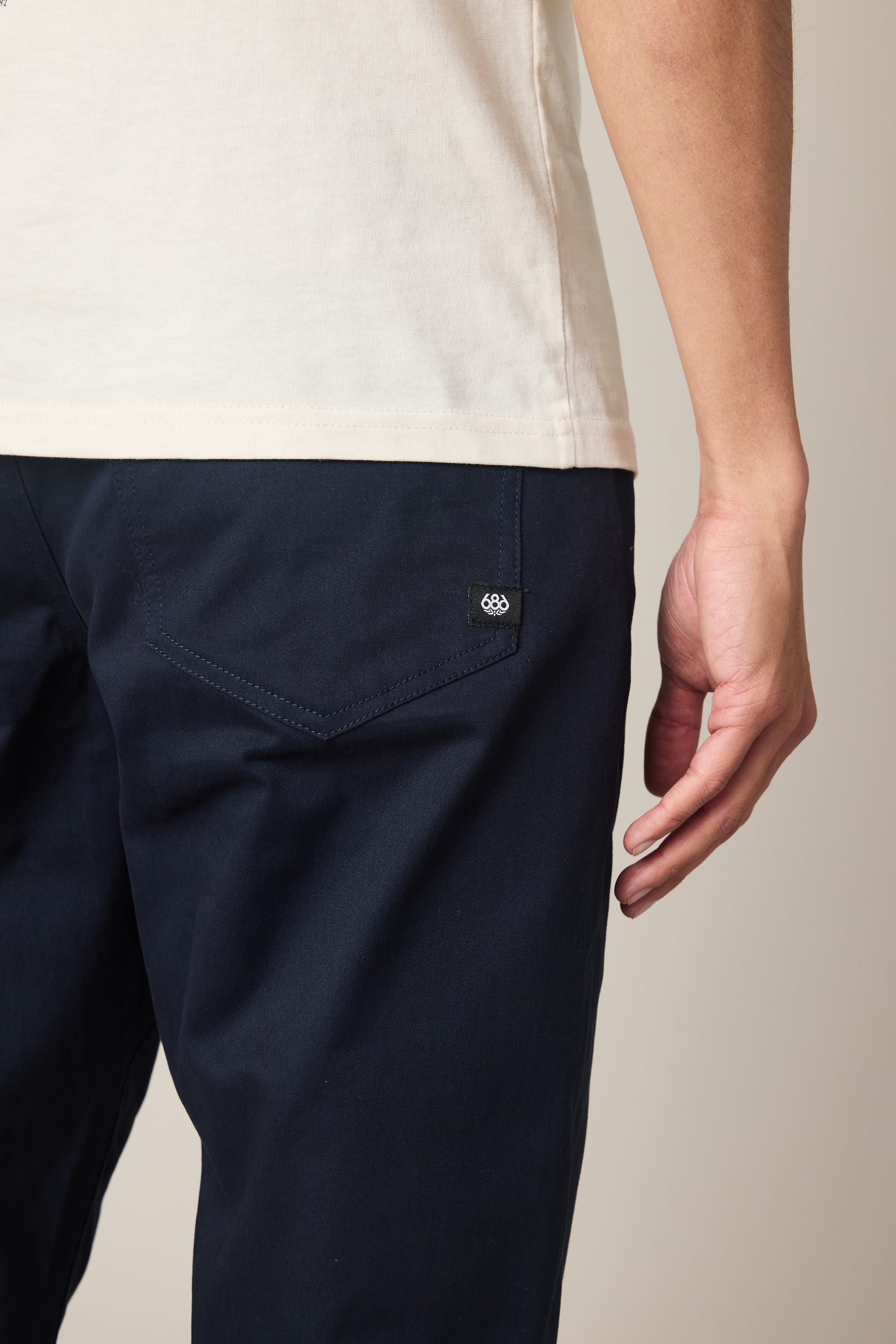 A person is shown from the back and side wearing a white t-shirt and 686 Men's Everywhere® Standard Pant - Slim Fit in dark blue, with their right hand by their hip. The pants have a buttoned back pocket for added style.