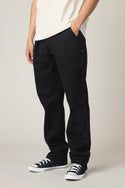 A person stands with one hand in their pocket, wearing 686 Men's Everywhere® Standard Pant - Relaxed Fit and a white t-shirt, along with black-and-white sneakers against a neutral background. The person's face is not visible.