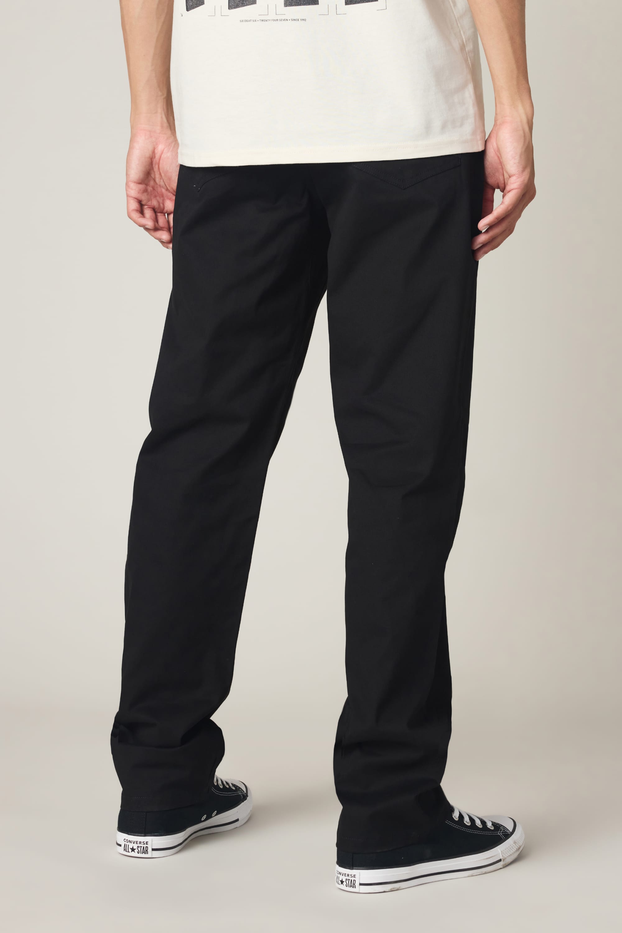A person stands facing away from the camera, wearing 686 Men's Everywhere® Standard Pant - Relaxed Fit by 686, black-and-white Converse sneakers, and a white T-shirt against a plain background.