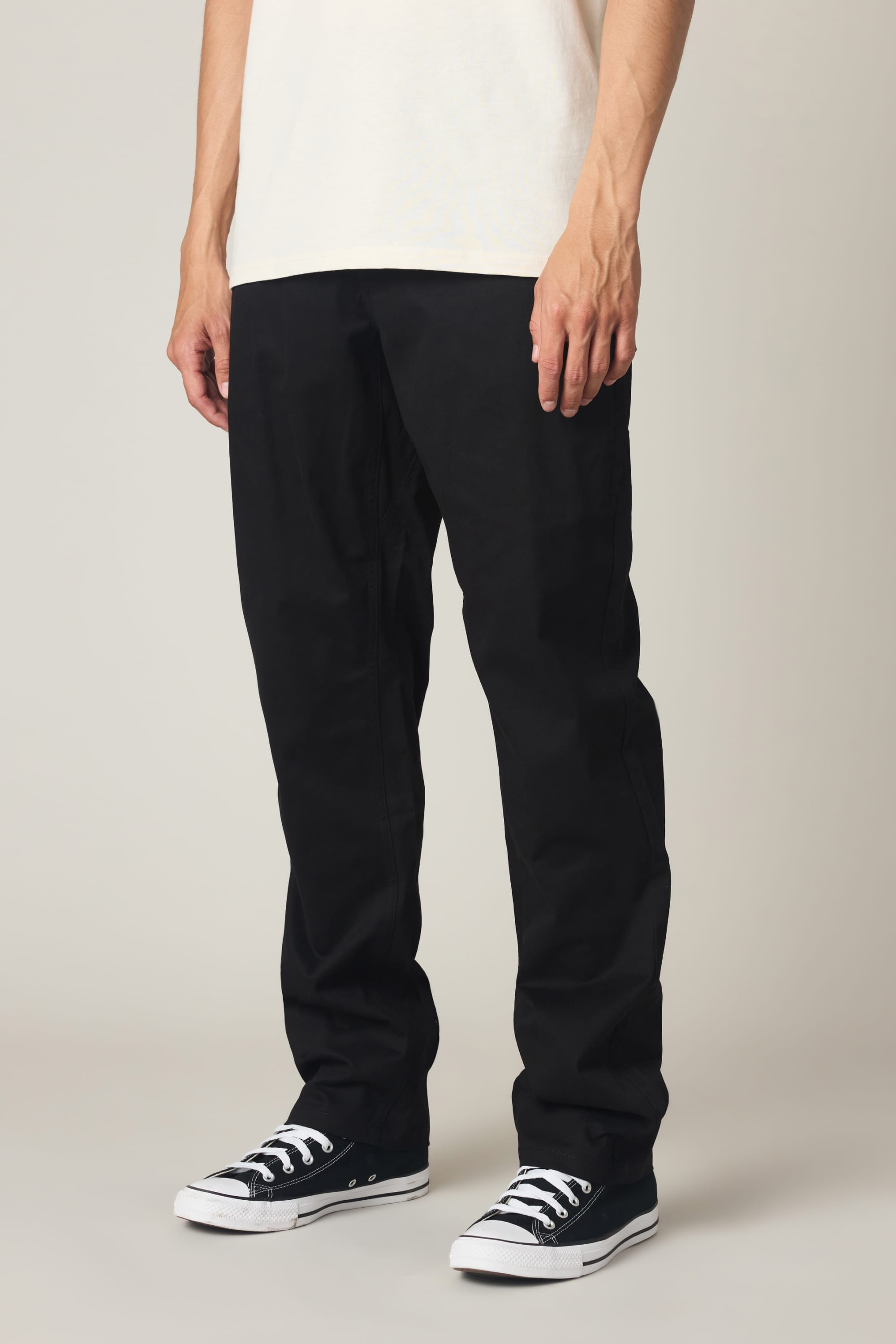 A person stands against a plain light background, wearing a white shirt, black low-top sneakers, and 686 Men's Everywhere® Standard Pant - Relaxed Fit by 686. The image is cropped from the shoulders down.