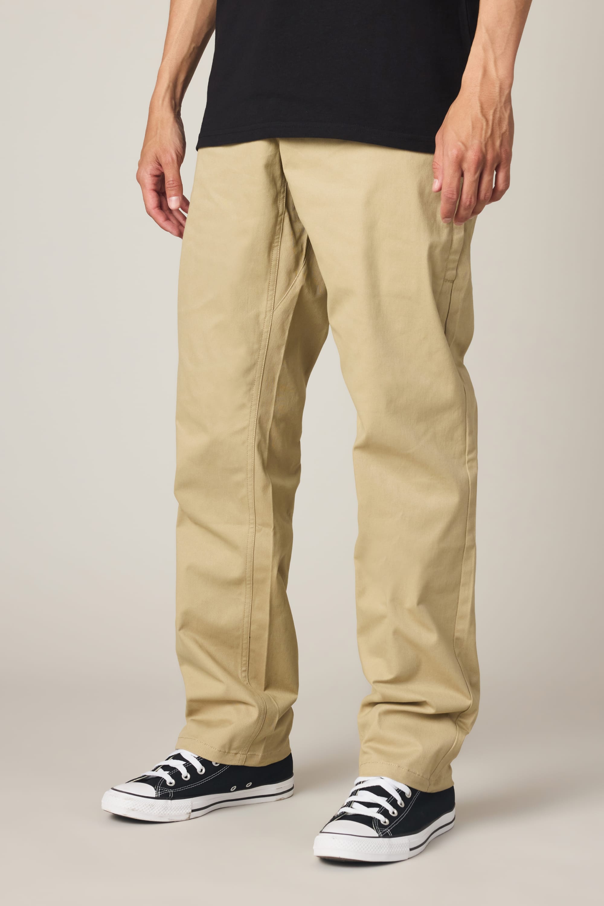 A person wears 686 Men's Everywhere® Standard Pant - Relaxed Fit in light khaki, a black t-shirt, and black low-top sneakers with white soles, standing against a plain background. The image is cropped at the waist.