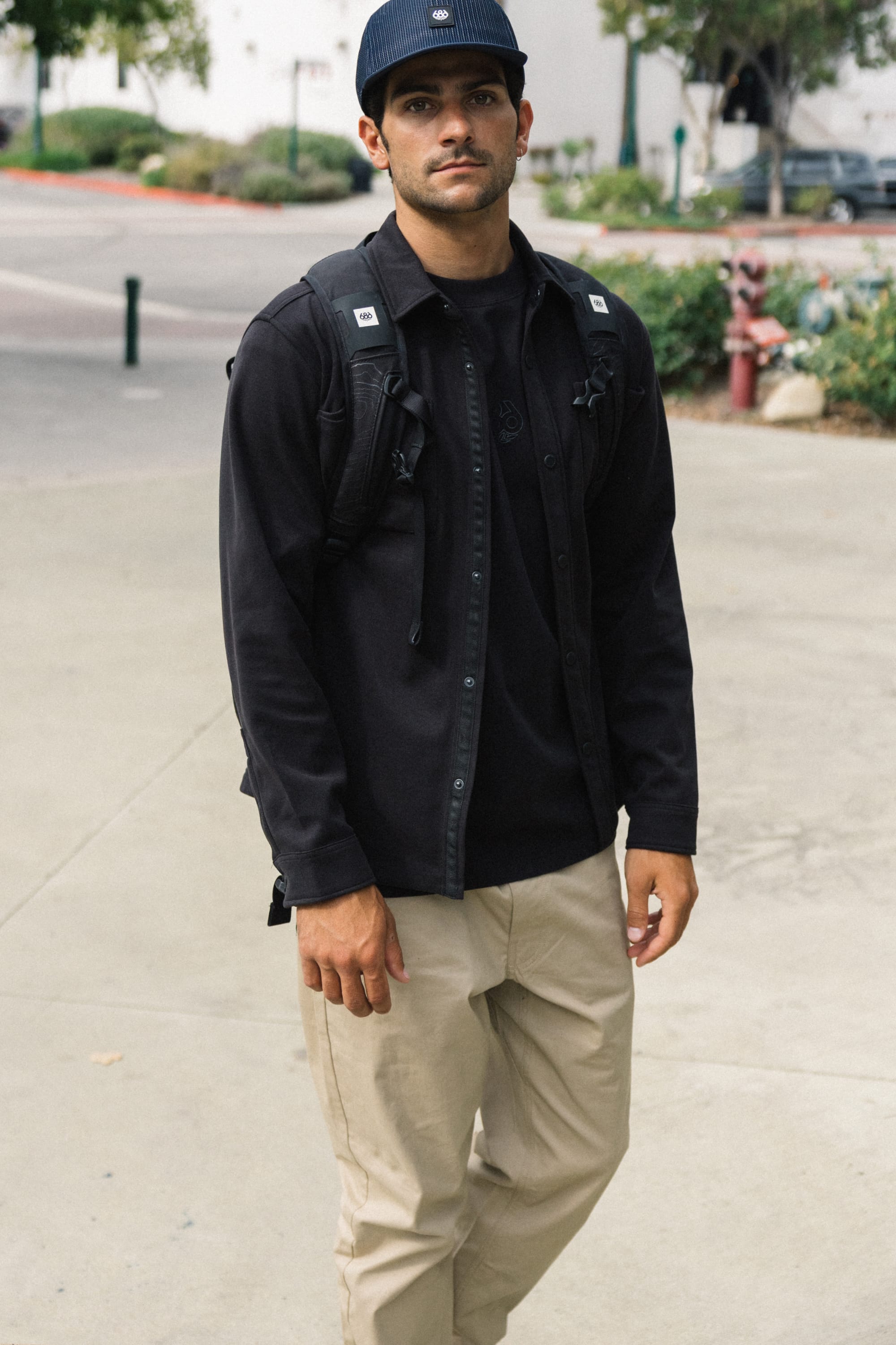 A man wearing a navy cap, black shirt, black backpack, and 686 Men's Everywhere® Standard Pant - Relaxed Fit by 686 stands on a sidewalk outdoors, looking at the camera.