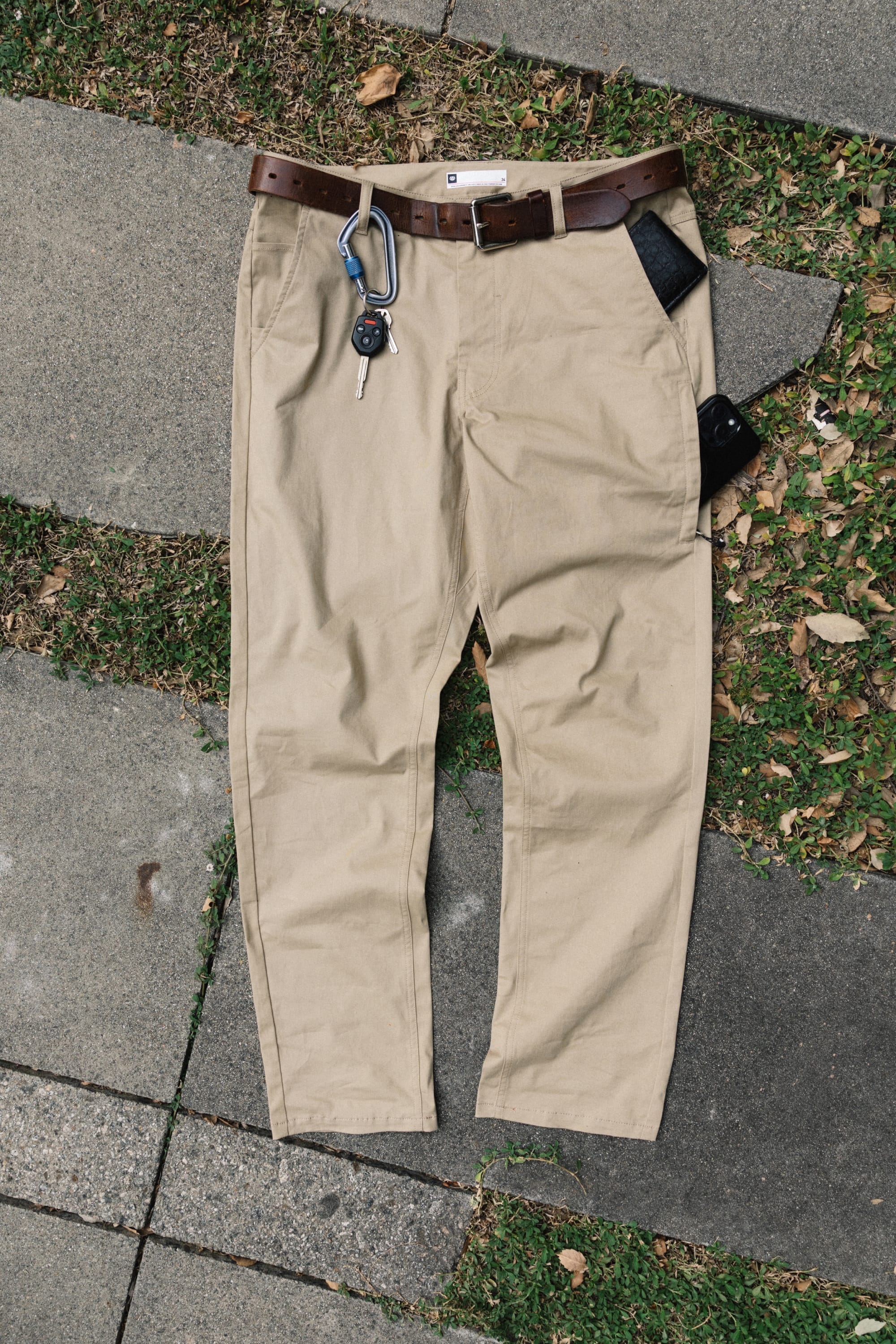 The 686 Men's Everywhere® Standard Pant - Relaxed Fit by 686 in beige is displayed with a brown belt, keys on a carabiner, and two black wallets on a stone and grass surface scattered with leaves.