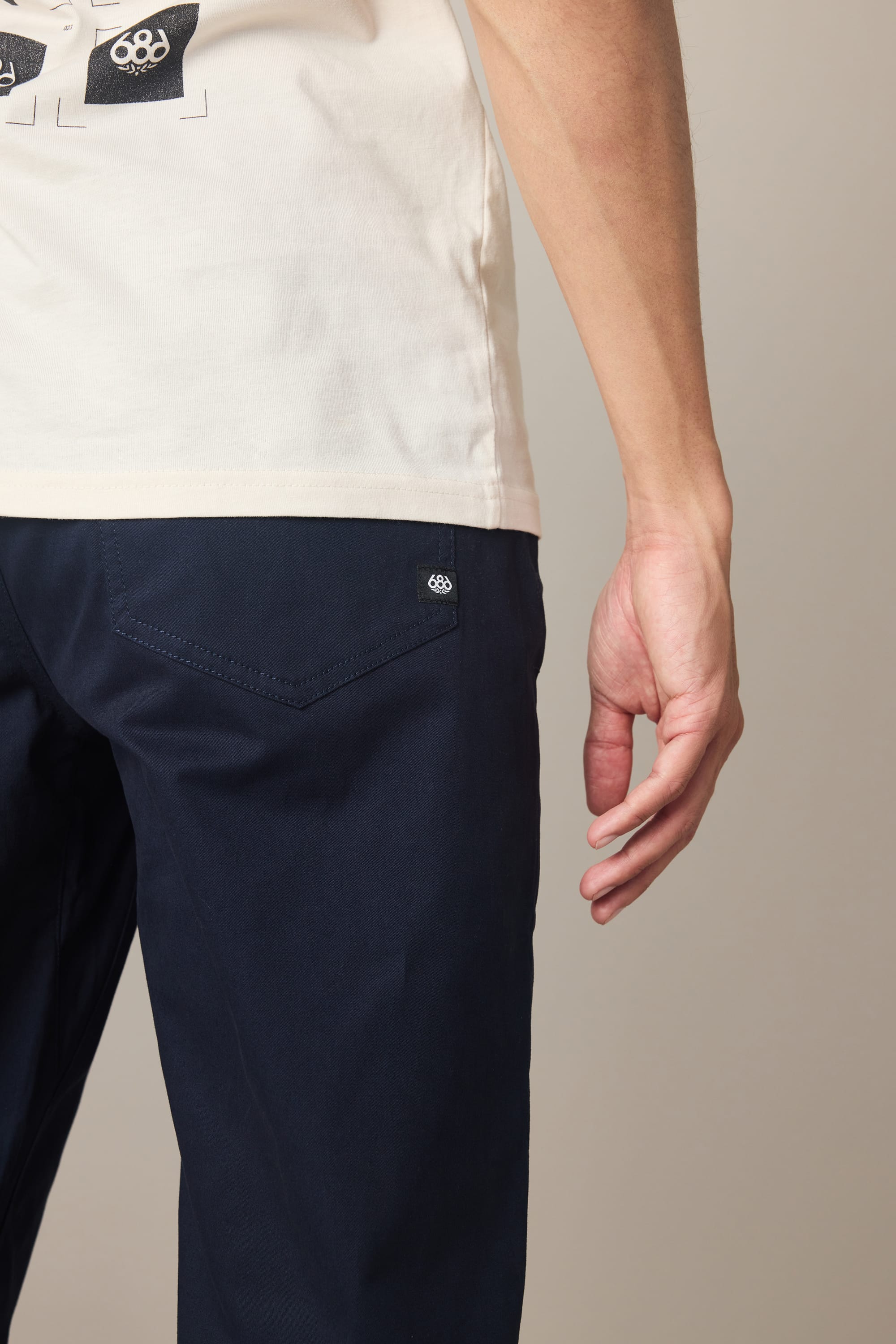 A person is shown from the waist down, standing sideways with one arm relaxed by their side while wearing a pair of 686 Men's Everywhere® Standard Pant - Relaxed Fit in navy blue and a white t-shirt.