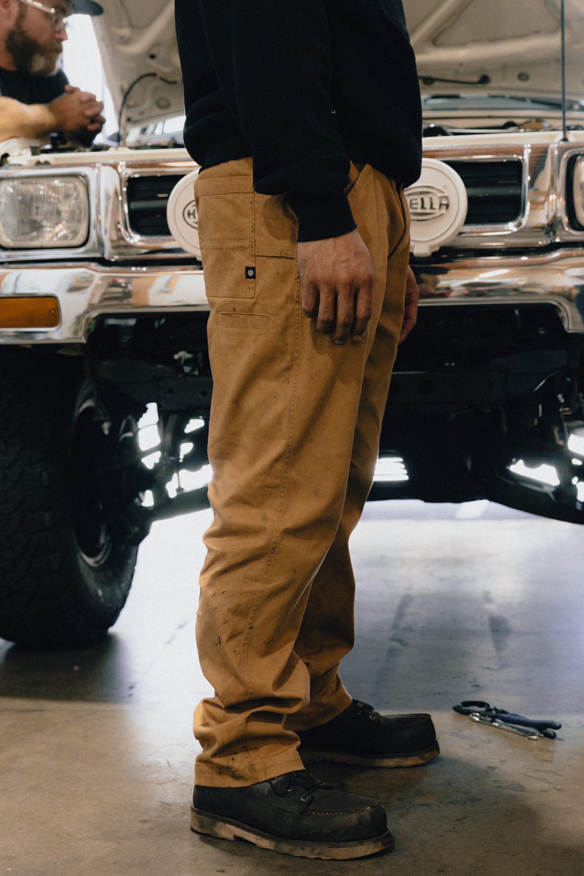 A person wearing the 686 Men's Unwork® Tradesman Pant - Relaxed Fit and dark shoes stands beside a vehicle with its hood open in a garage. Tools are scattered on the floor, and another individual is visible in the background near the car’s grill.