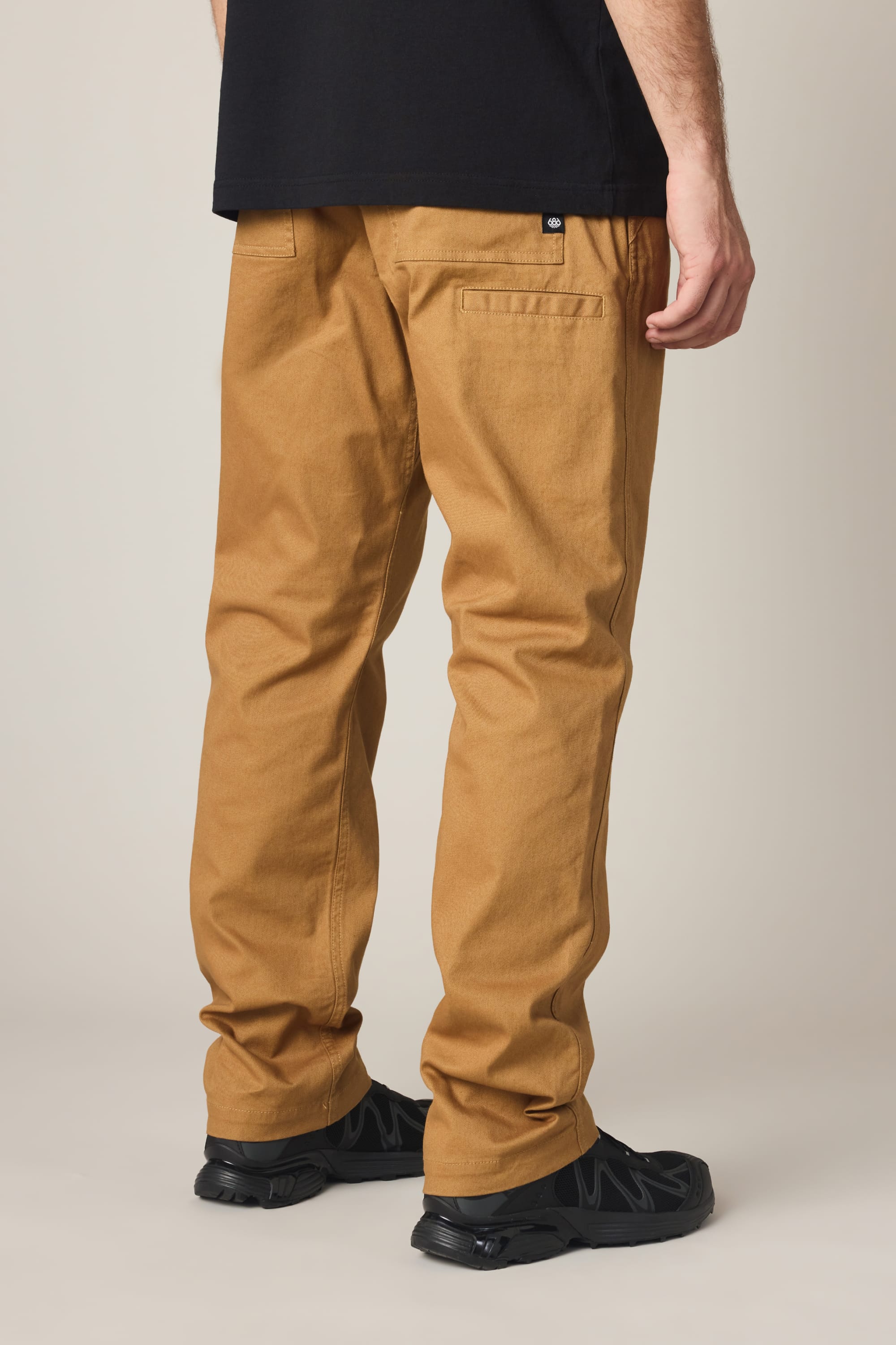 A person wearing the 686 Men's Unwork® Tradesman Pant - Relaxed Fit in tan, paired with black shoes and a black shirt, stands with their back turned. Only the lower half of the body is visible.