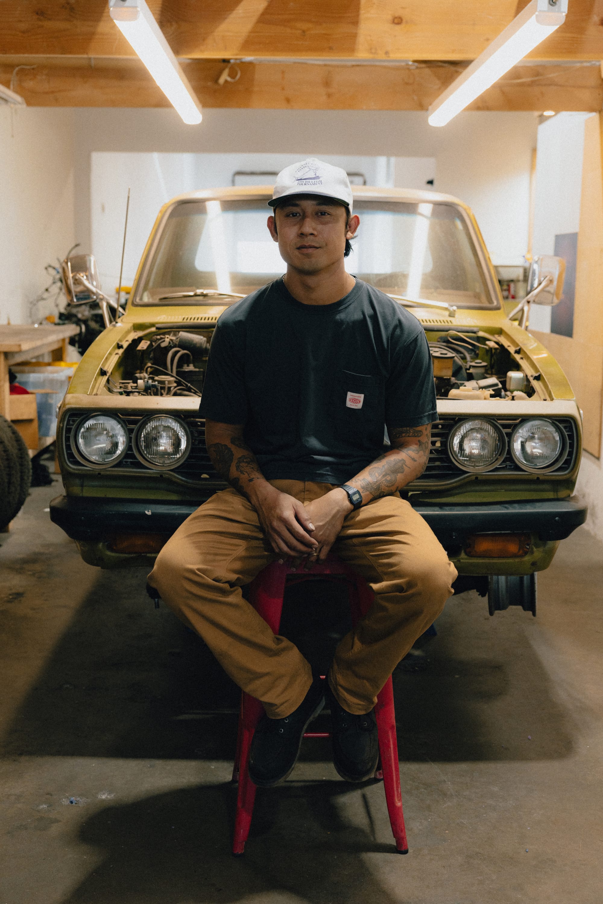 A man wearing a cap and casual clothes, including the 686 Men's Unwork® Tradesman Pant - Relaxed Fit by 686, sits on a red stool in front of a vintage yellow car in a garage, surrounded by tools and car parts.