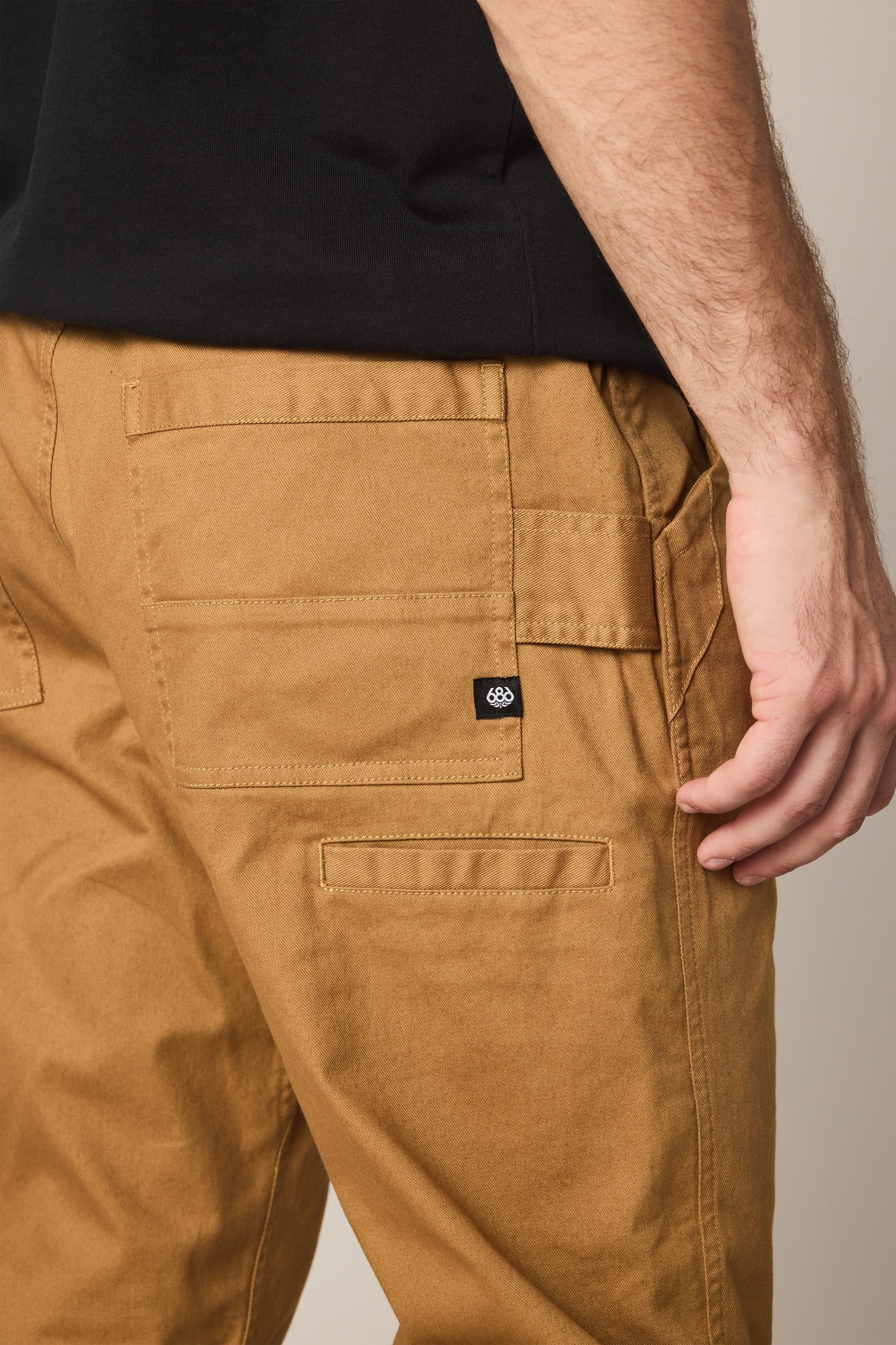 A person is shown from the waist down wearing 686 Men's Unwork® Tradesman Pant - Relaxed Fit in tan, paired with a black shirt, with their right hand resting near the pant's side pocket.
