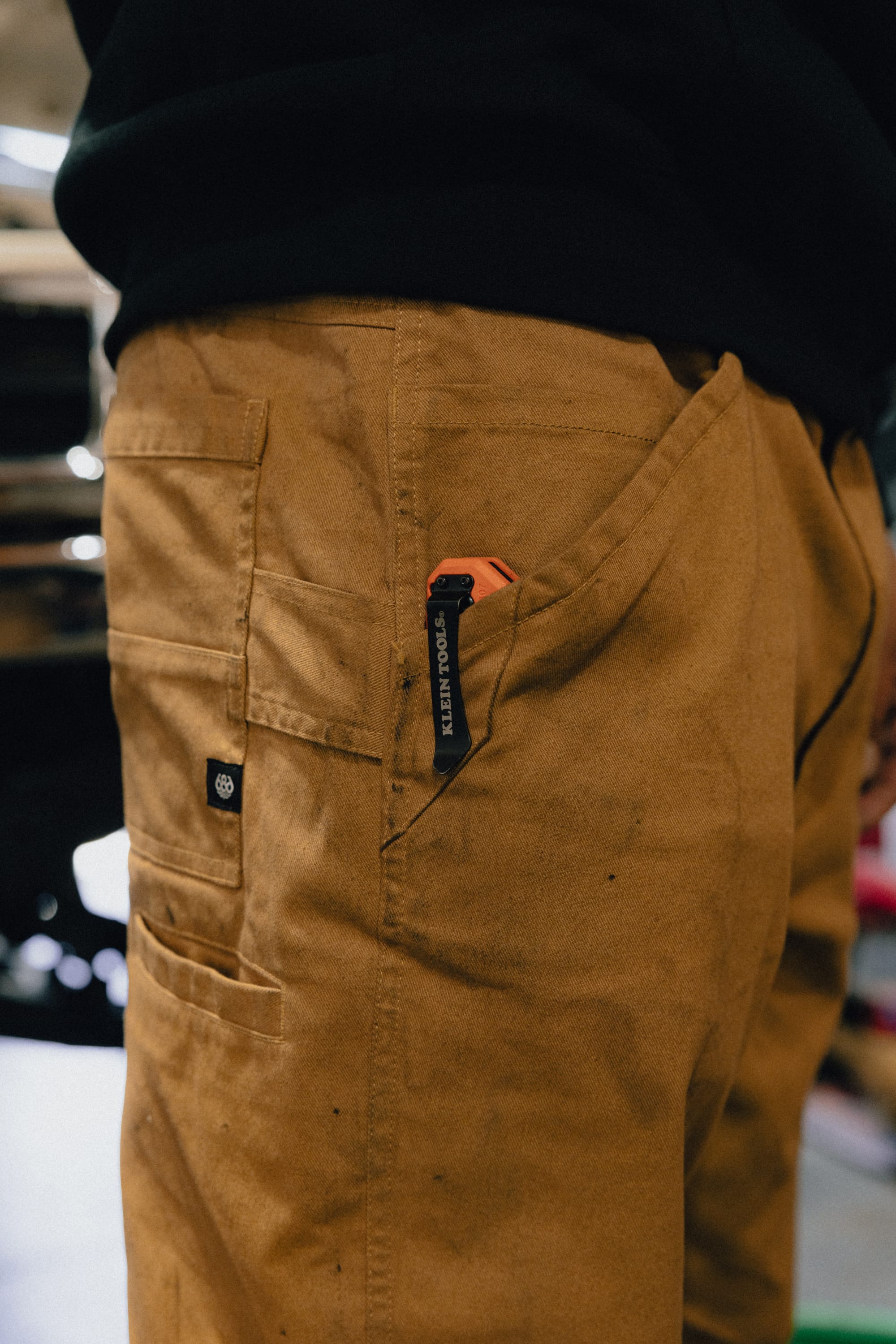 A person wearing tan 686 Men's Unwork® Tradesman Pant - Relaxed Fit has a folding utility knife clipped to their back pocket. The black knife has a visible HEALTH TOPIC label on the clip.