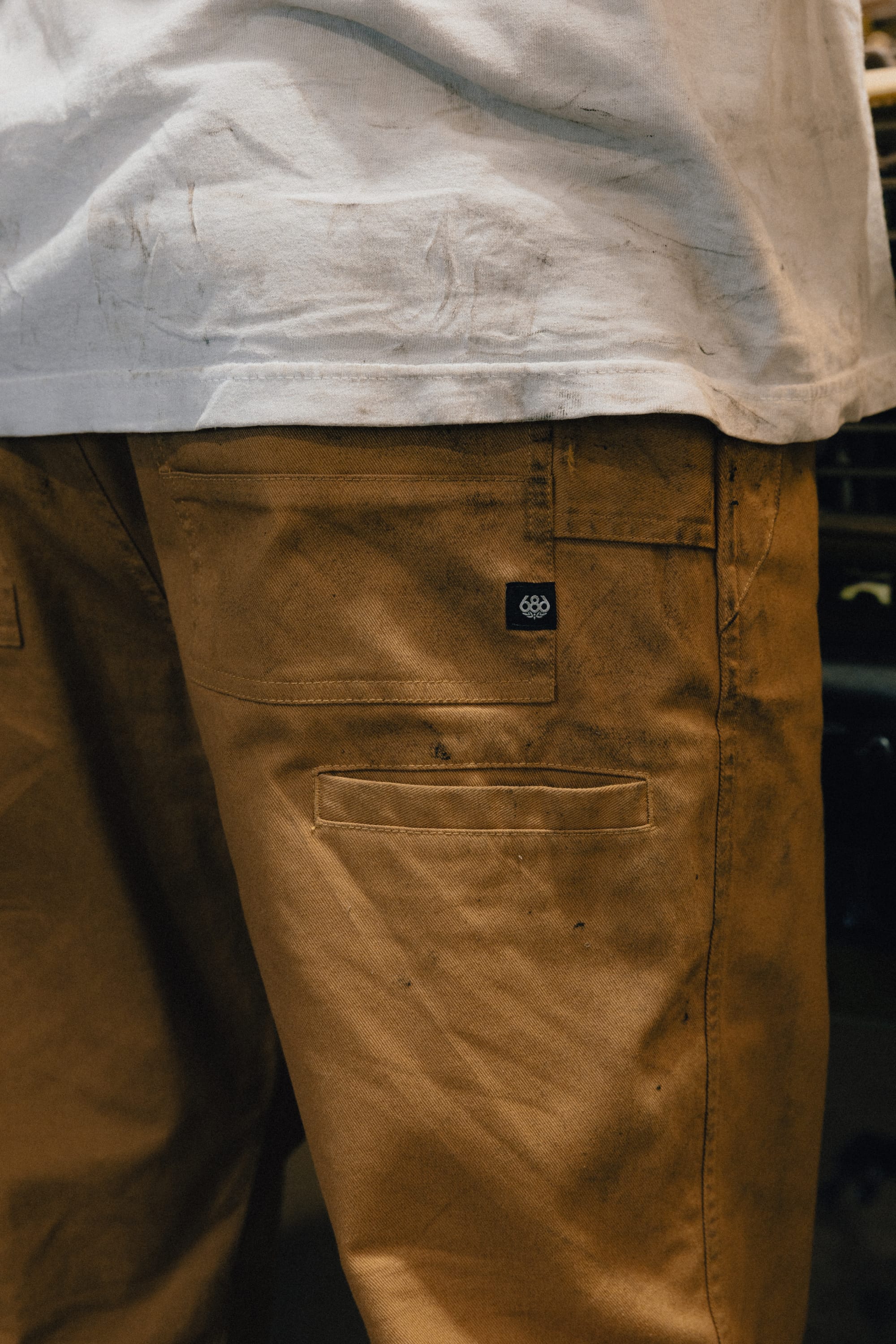 A person wearing a dirty white shirt and stained brown 686 Men's Unwork® Tradesman Pant - Relaxed Fit stands with their back to the camera. A small black tag with a white 686 logo is visible above the pocket on the pants.