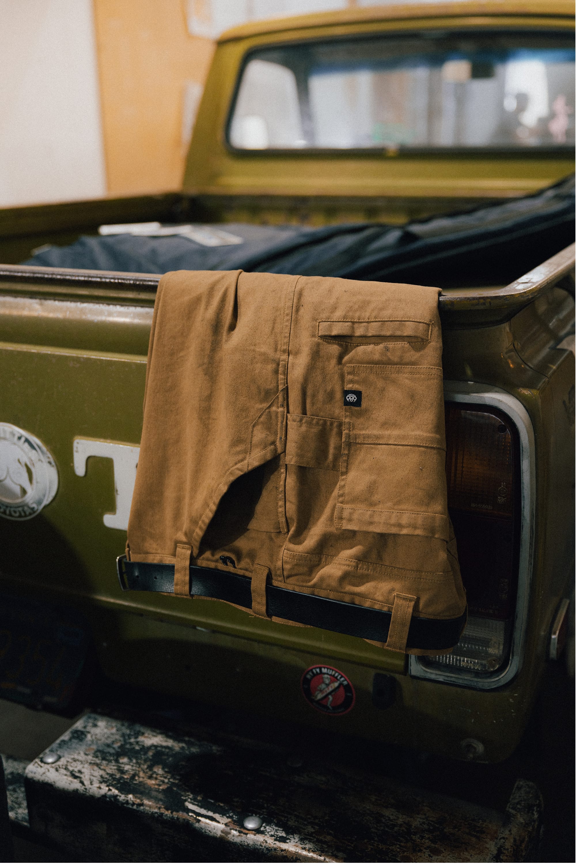 A pair of 686 Men's Unwork® Tradesman Pant - Relaxed Fit in brown is draped over the tailgate of an olive green pickup truck, which appears old with visible rust and weathering.