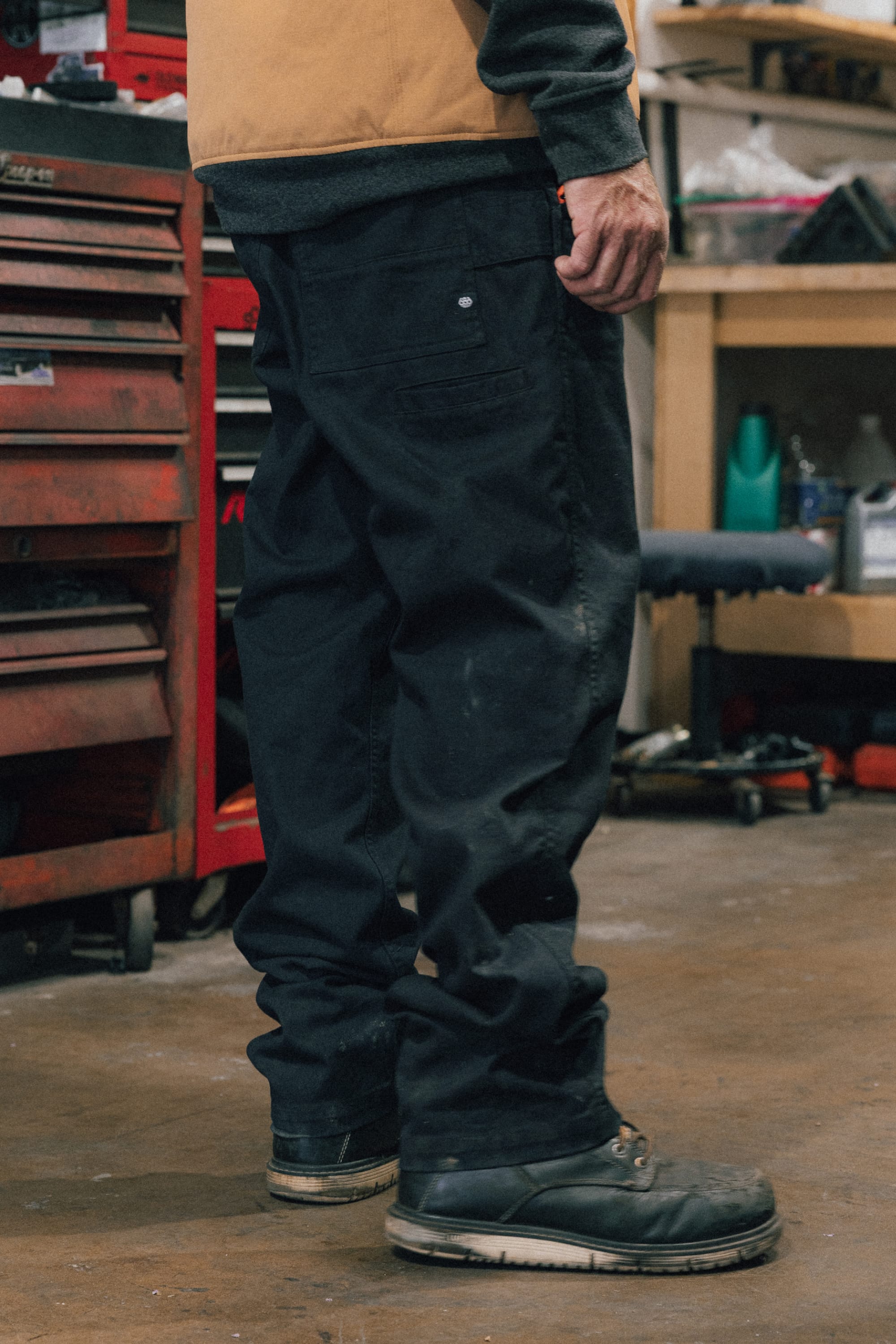 A person wearing the 686 Men's Unwork® Tradesman Pant - Relaxed Fit stands in a workshop with a red tool chest and shelves in the background.