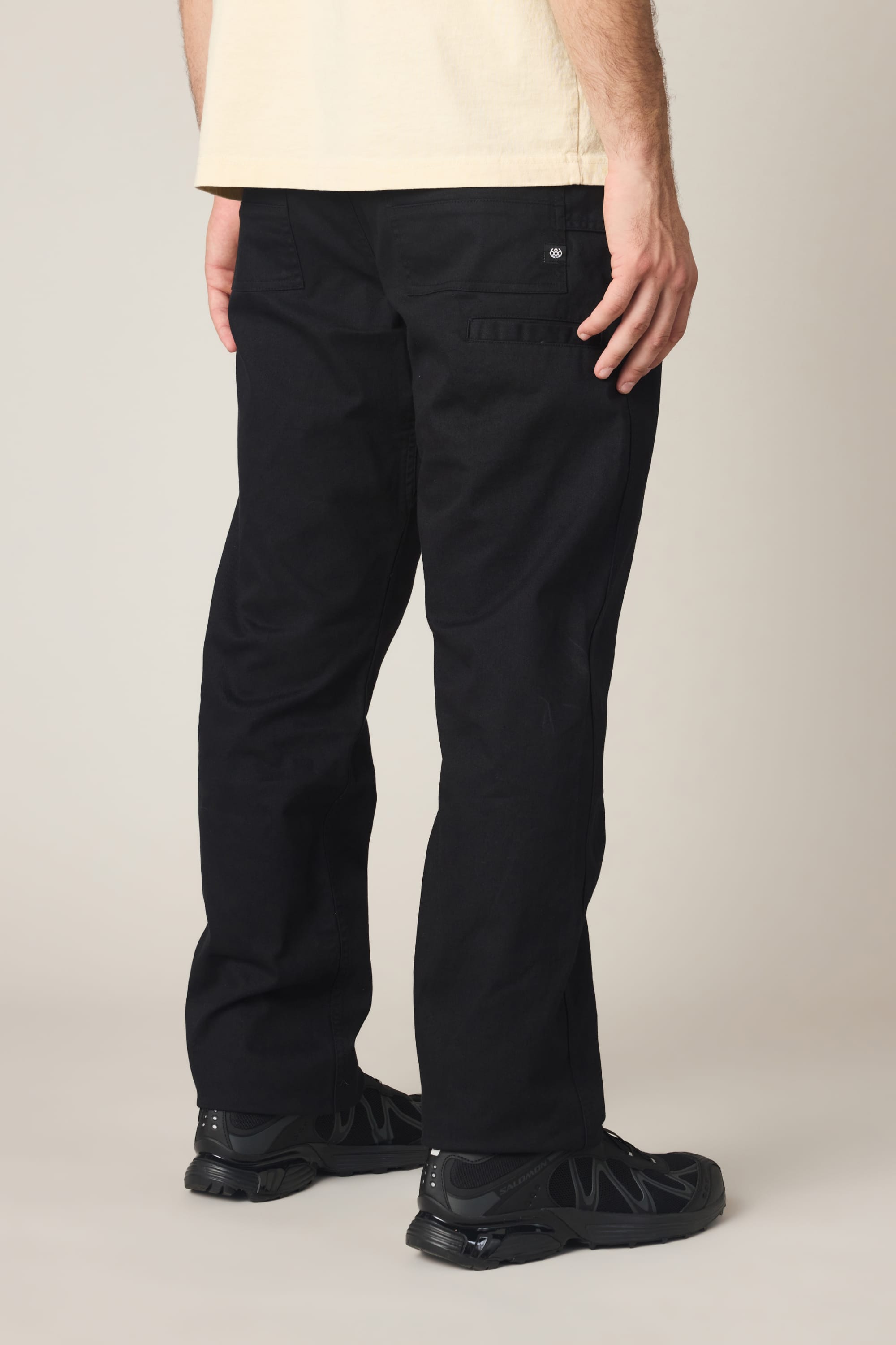 A person stands facing away, wearing a beige T-shirt and black sneakers, showcasing the 686 Men's Unwork® Tradesman Pant - Relaxed Fit by 686, featuring a back pocket flap, against a plain background.