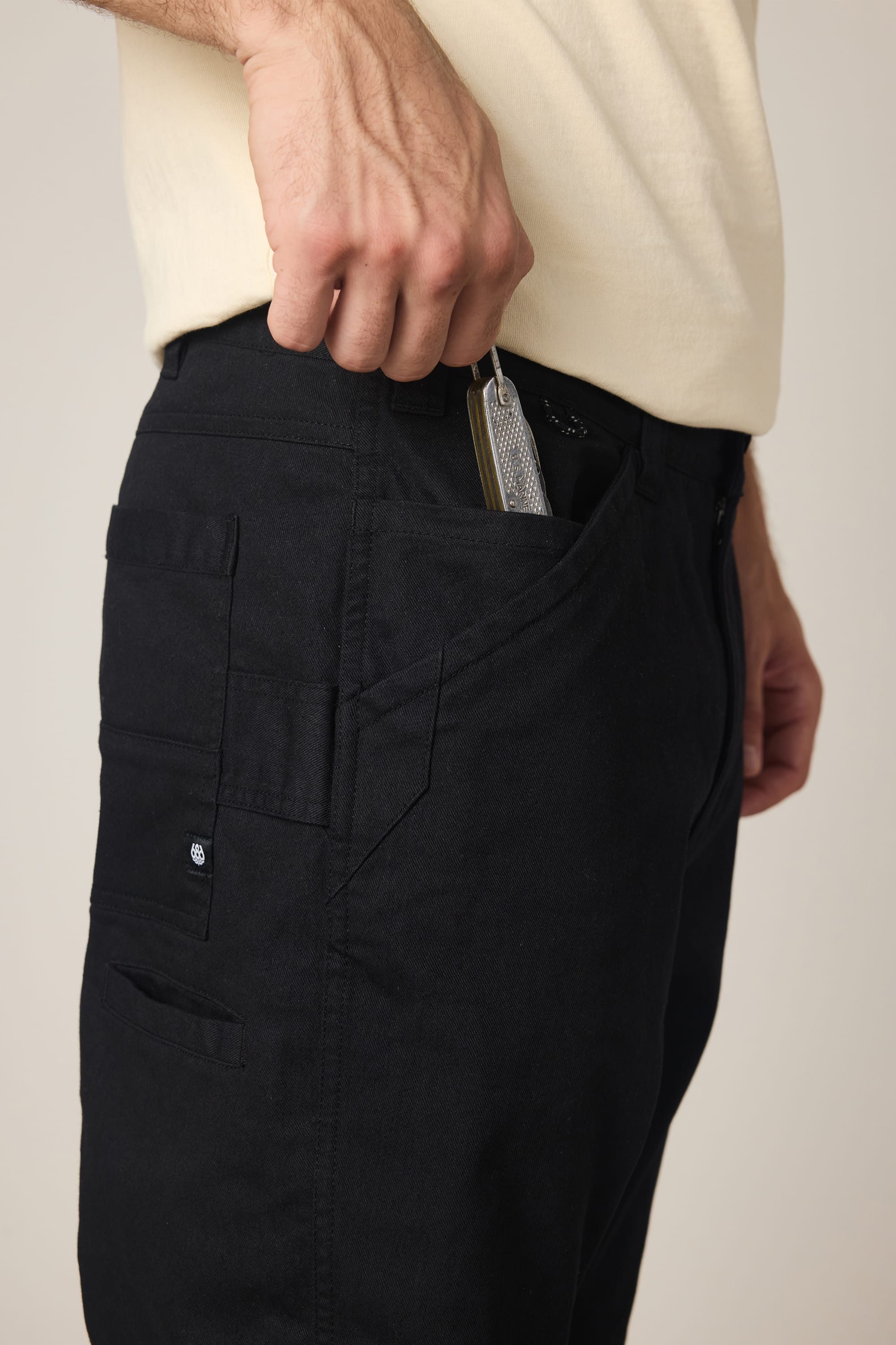 A person in a light-colored shirt and 686 Men's Unwork® Tradesman Pant - Relaxed Fit places a smartphone into the front pocket of their pants with their right hand.