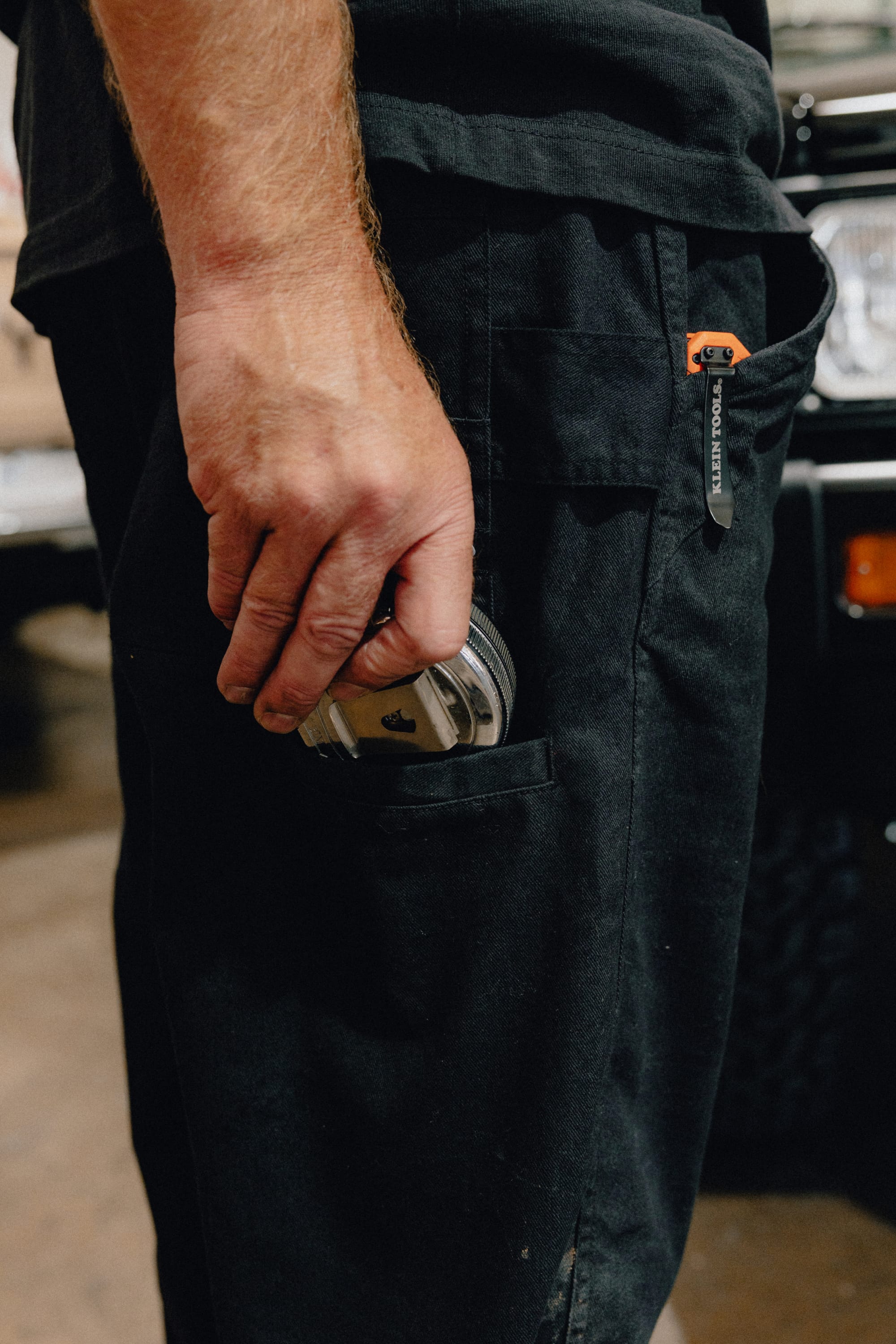 A person wearing the 686 Men's Unwork® Tradesman Pant - Relaxed Fit is holding a round metal object next to their side pocket. The background includes part of a vehicle and an indoor setting.