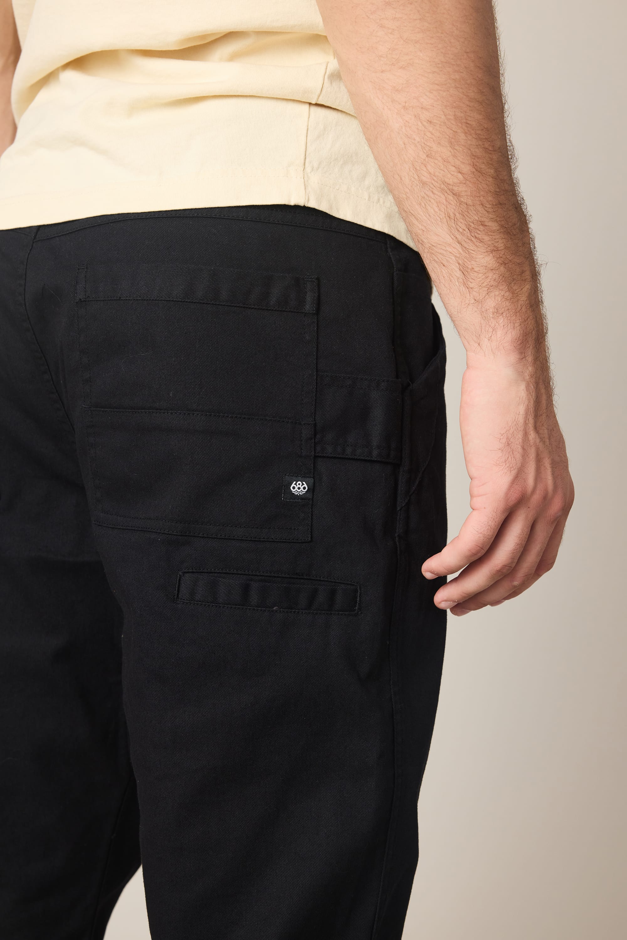 A person wearing the 686 Men's Unwork® Tradesman Pant - Relaxed Fit in black, shown from the waist down and behind, with their right hand relaxed by their side and a back pocket featuring a small stitched 686 logo.