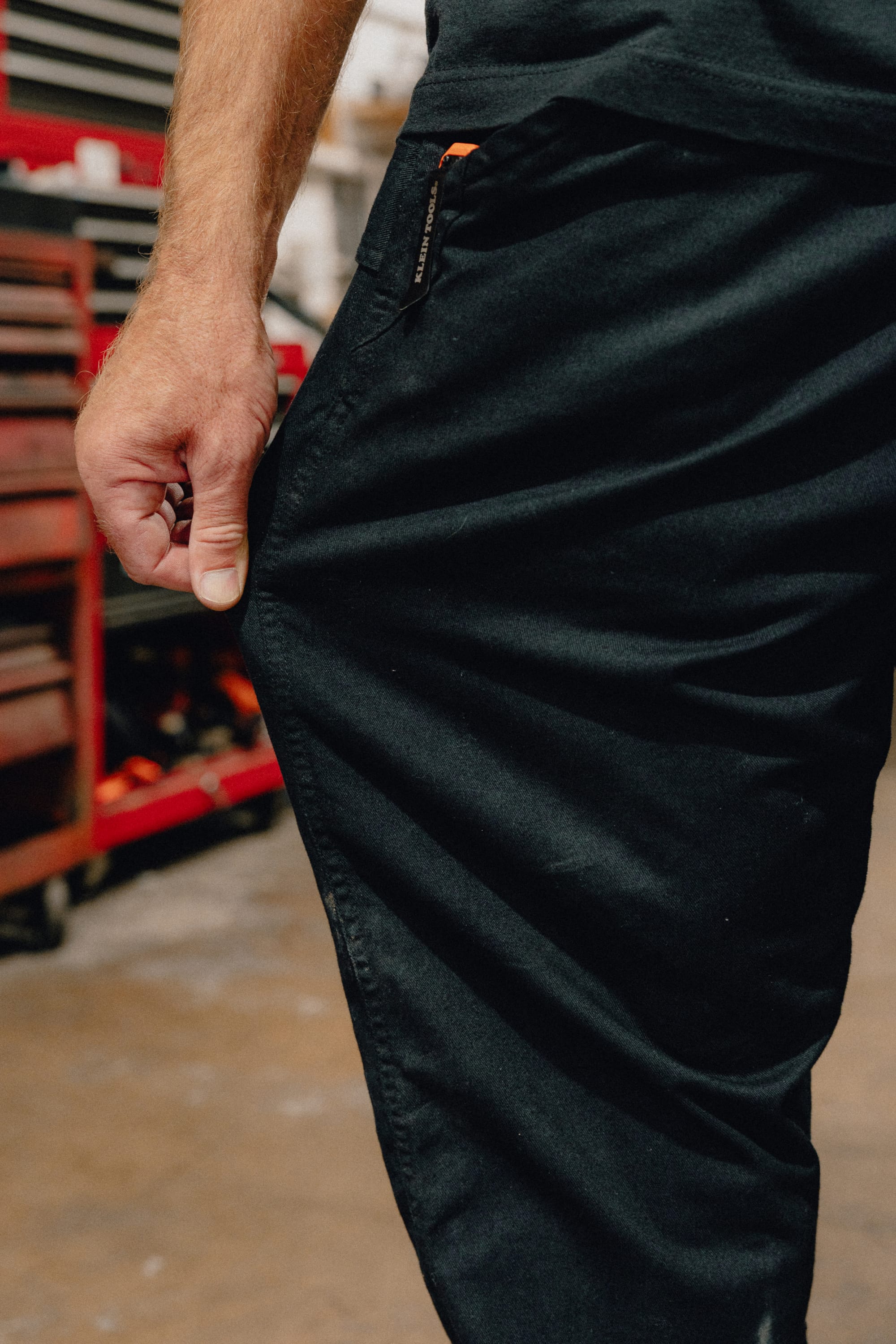 A person wearing the 686 Men's Unwork® Tradesman Pant - Relaxed Fit stands indoors, pulling at their pocket with their right hand. The background features a red tool chest and workshop equipment in soft focus.
