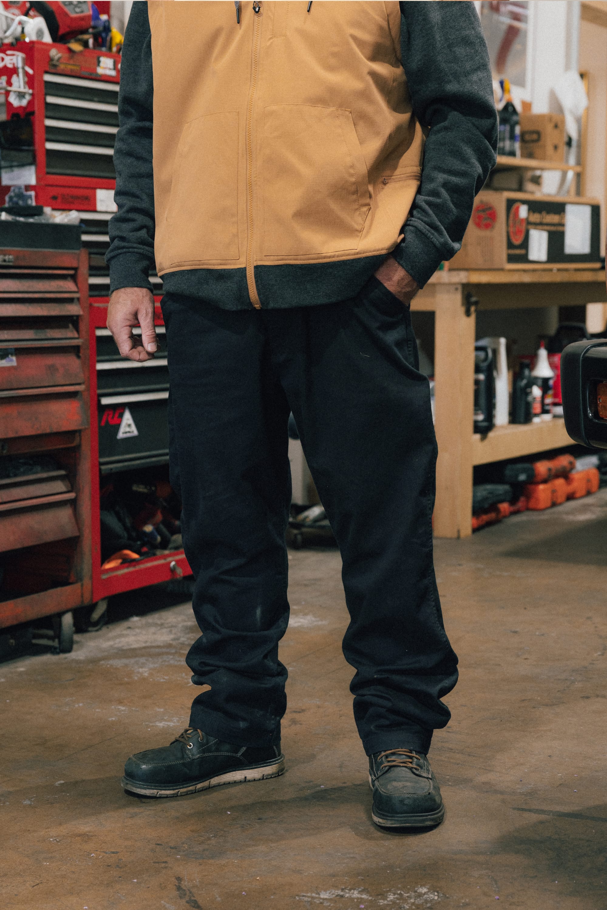A person stands in a garage with tools and toolboxes in the background, wearing a tan vest, gray sweatshirt, and 686 Men's Unwork® Tradesman Pant - Relaxed Fit by 686, paired with work boots. Their face is not visible.