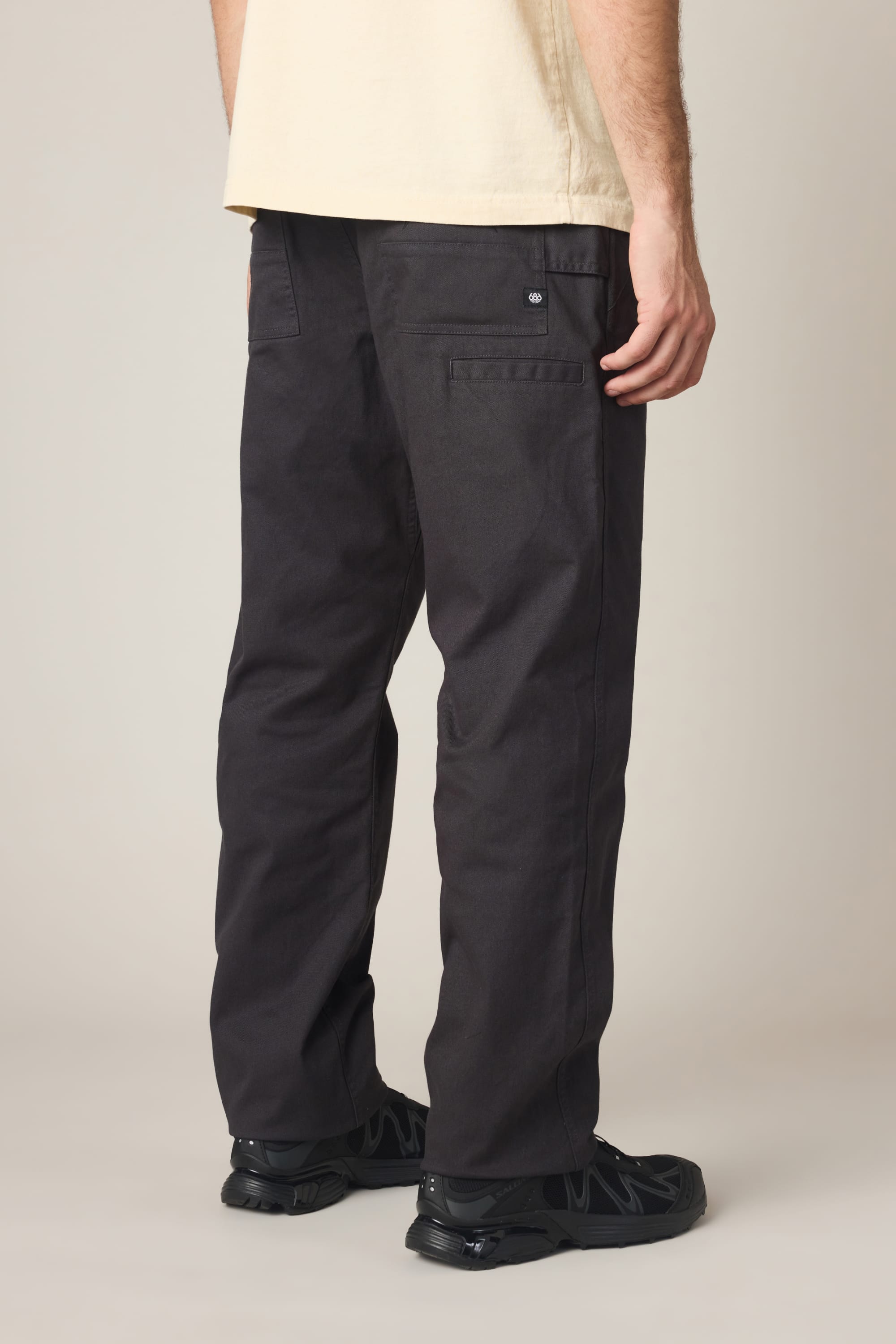 A person stands facing away, wearing a light beige T-shirt and the 686 Men's Unwork® Tradesman Pant - Relaxed Fit in dark gray, featuring back pockets and paired with black shoes, against a plain neutral background.