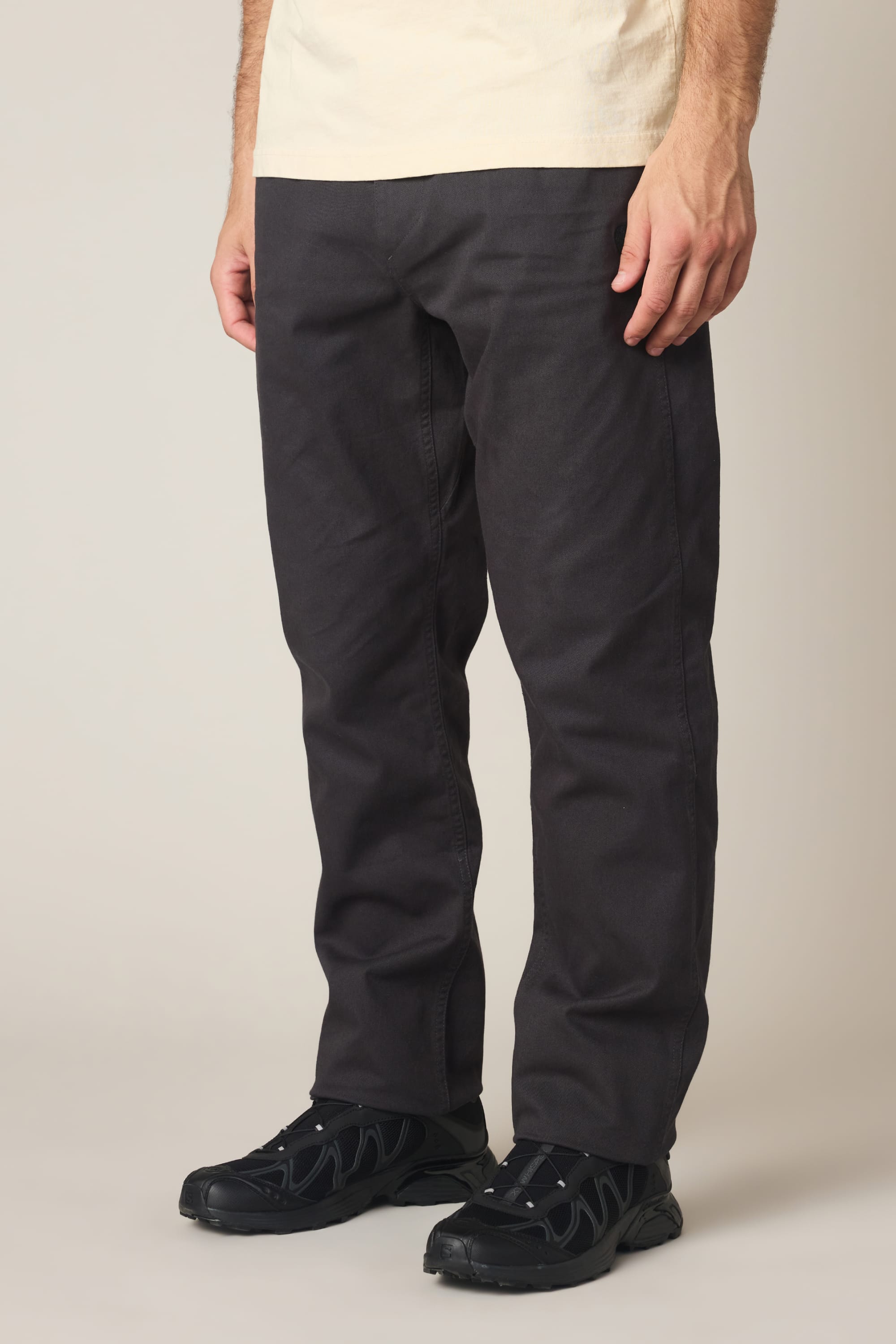 A person wearing a light beige shirt, 686 Men's Unwork® Tradesman Pant - Relaxed Fit in dark gray, and black sneakers stands against a plain light background; only the lower half of their body is visible.