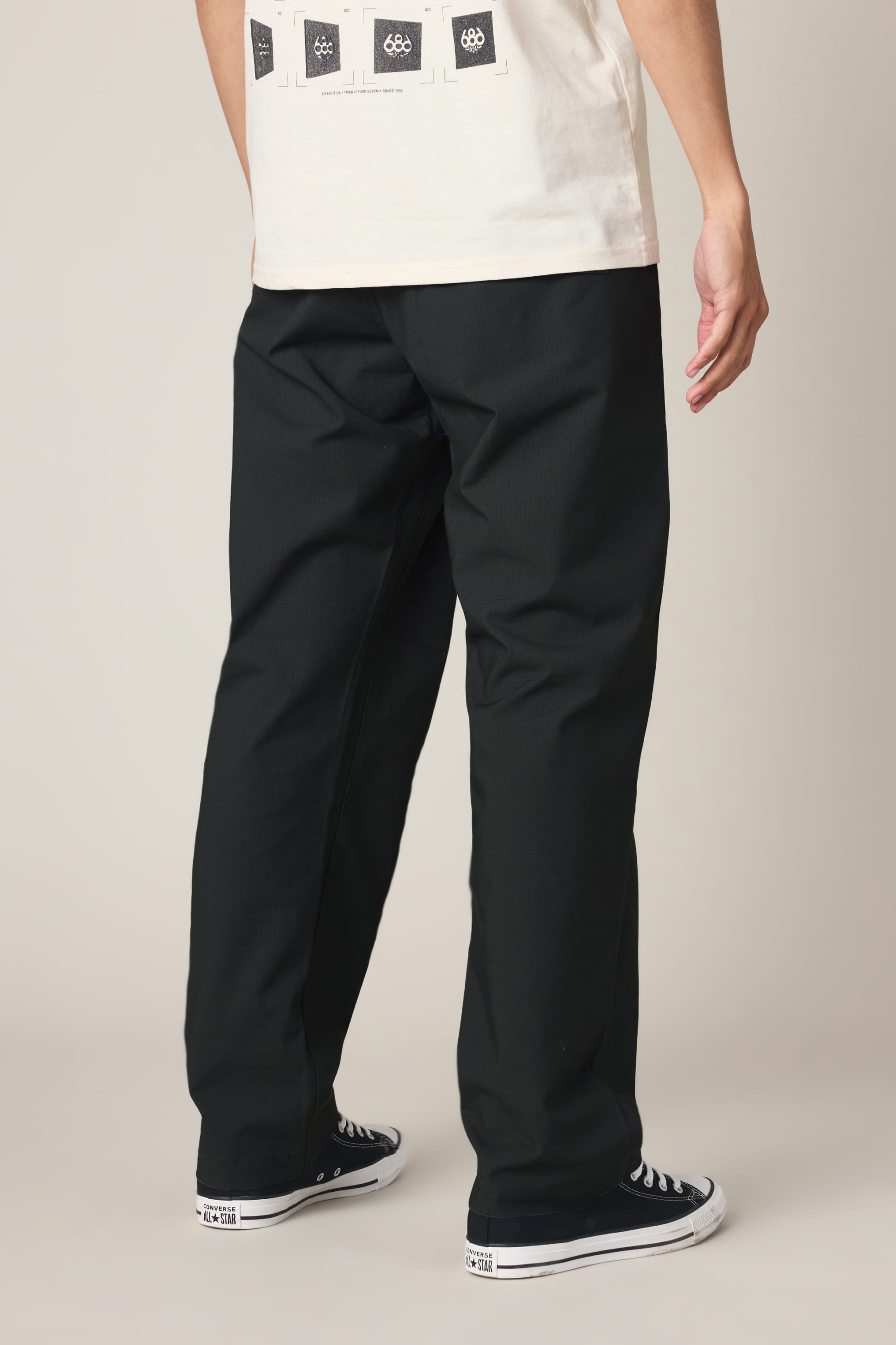 A person stands with their back turned, wearing 686 Men's Free Loader Pant - Loose Fit by 686, black and white sneakers, and a white T-shirt with graphics on the back.