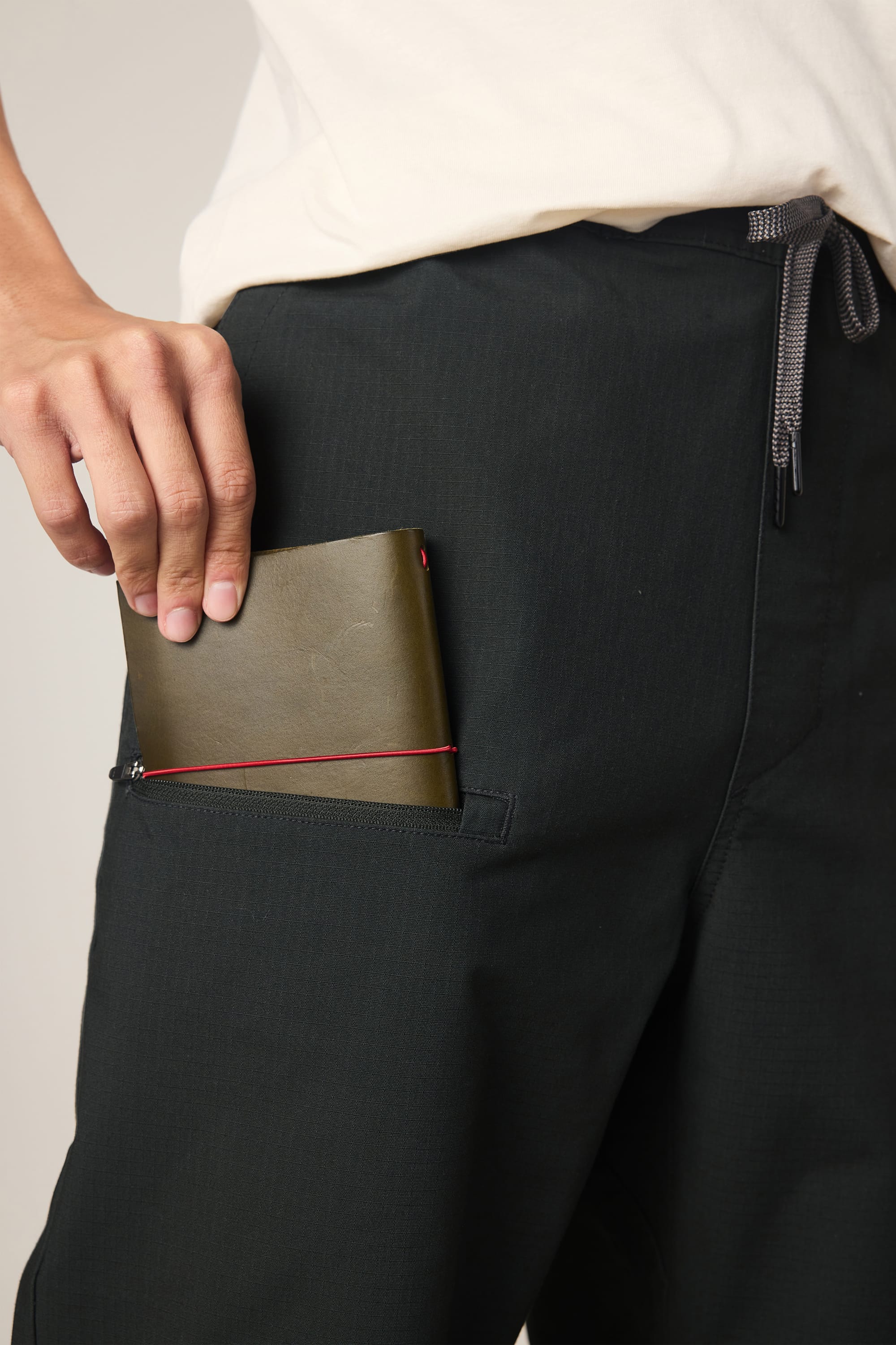A person wearing the 686 Men's Free Loader Pant - Loose Fit and a white shirt places a green notebook with a red elastic band into the pant’s zippered side pocket.