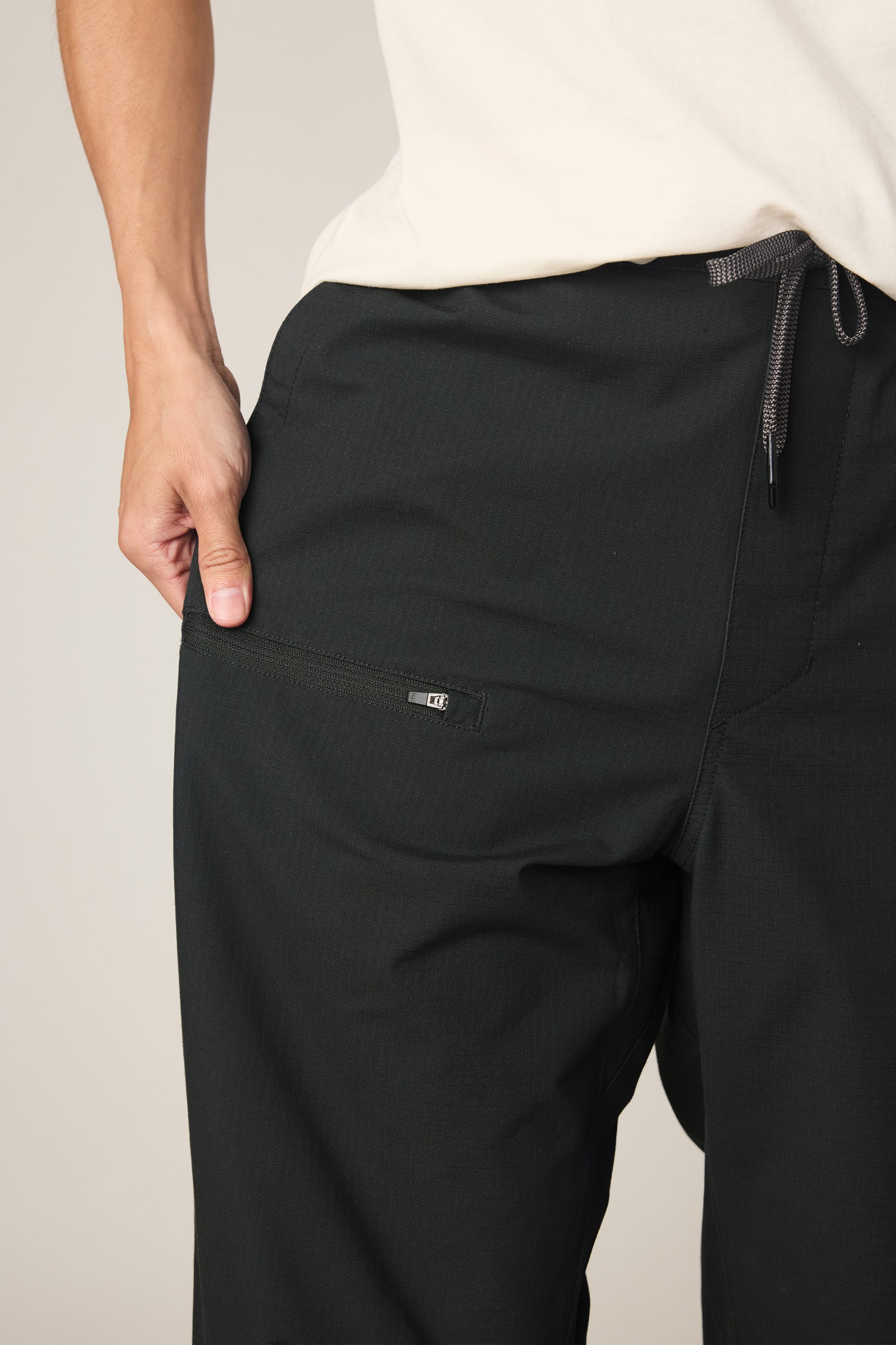 A person wearing the 686 Men's Free Loader Pant - Loose Fit in black by 686, paired with a white shirt, stands with one hand in their pocket. The nylon woven pant showcases a visible zipper on the pocket and a drawstring at the waist.