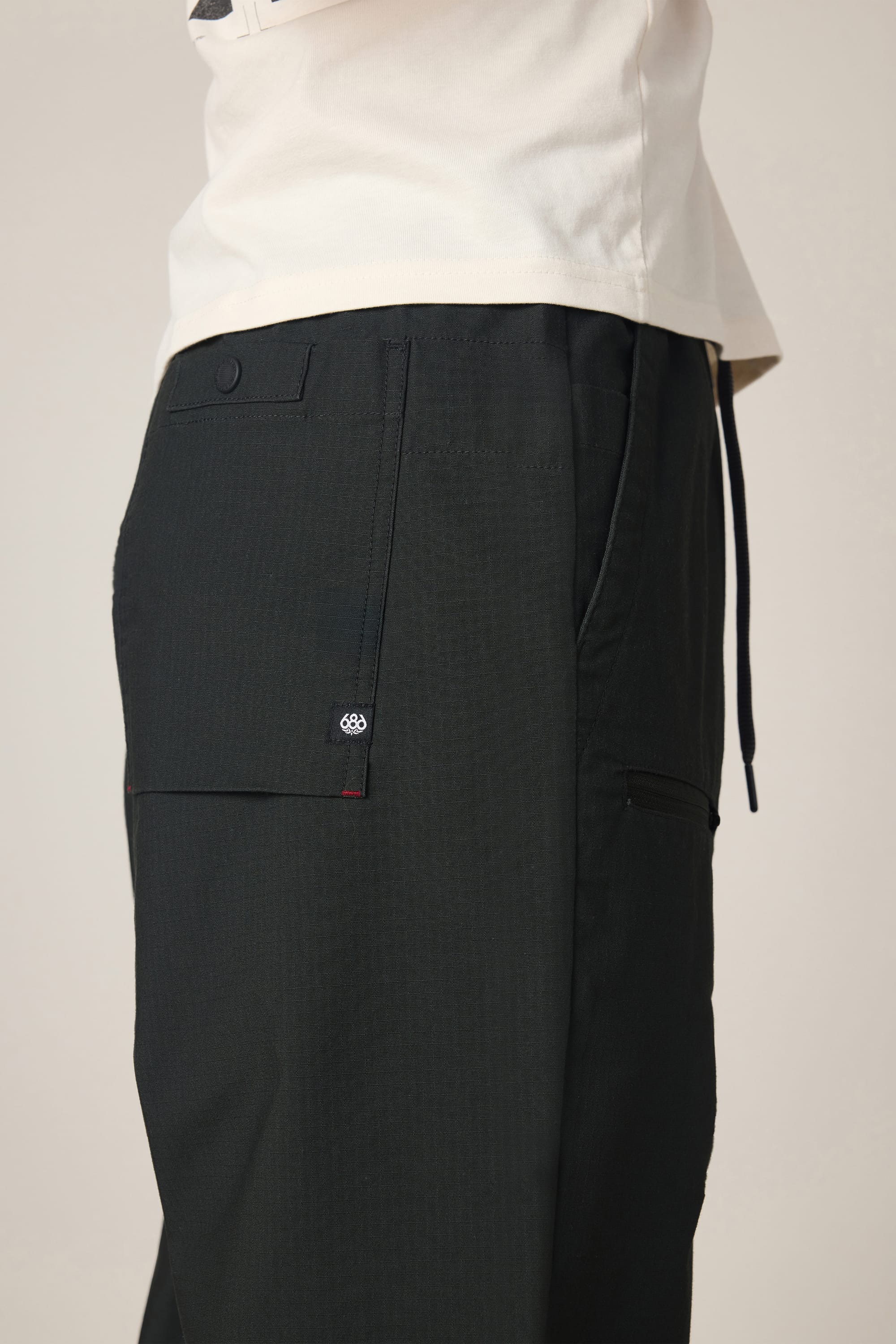 Side view of a person wearing 686 Men's Free Loader Pant - Loose Fit in black, paired with a white shirt. The image showcases the waist, upper leg, flap pocket detail, and part of the drawstring on these modern spandex pants by 686.