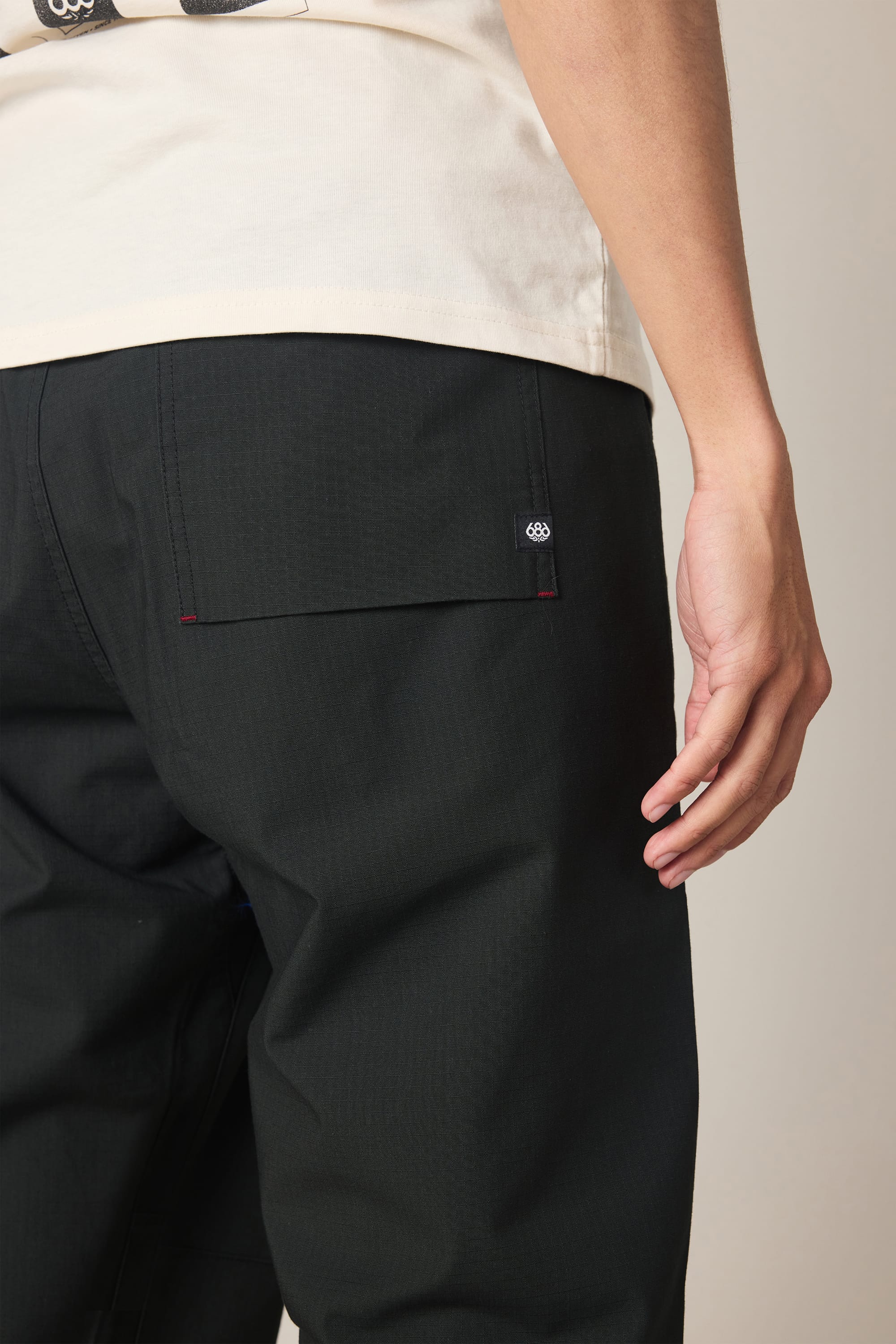 A person wearing the 686 Men's Free Loader Pant - Loose Fit in black, paired with a light shirt, is shown from the waist down and from the back, with their right hand relaxed by their side near the pocket.