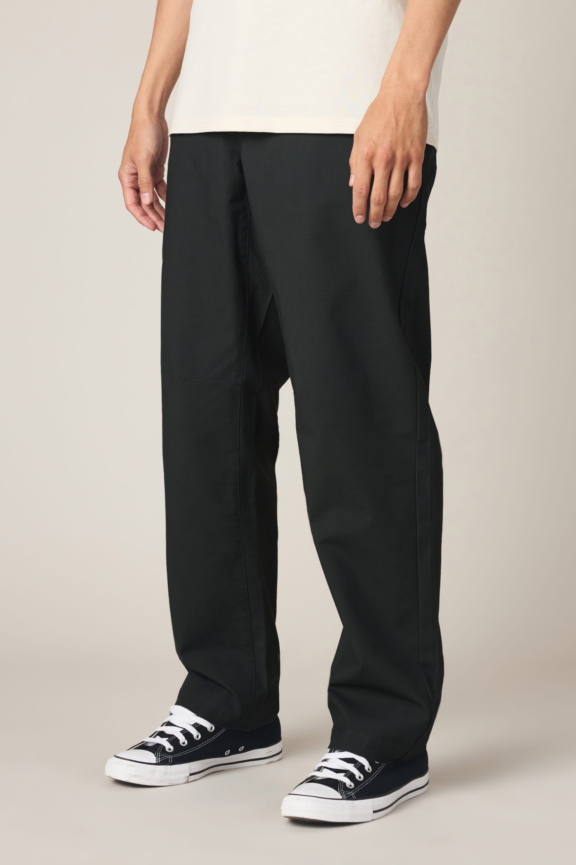 A person is shown from the waist down, standing against a plain light background, wearing the 686 Men's Free Loader Pant - Loose Fit by 686 in black, paired with a white shirt and black low-top sneakers with white laces.
