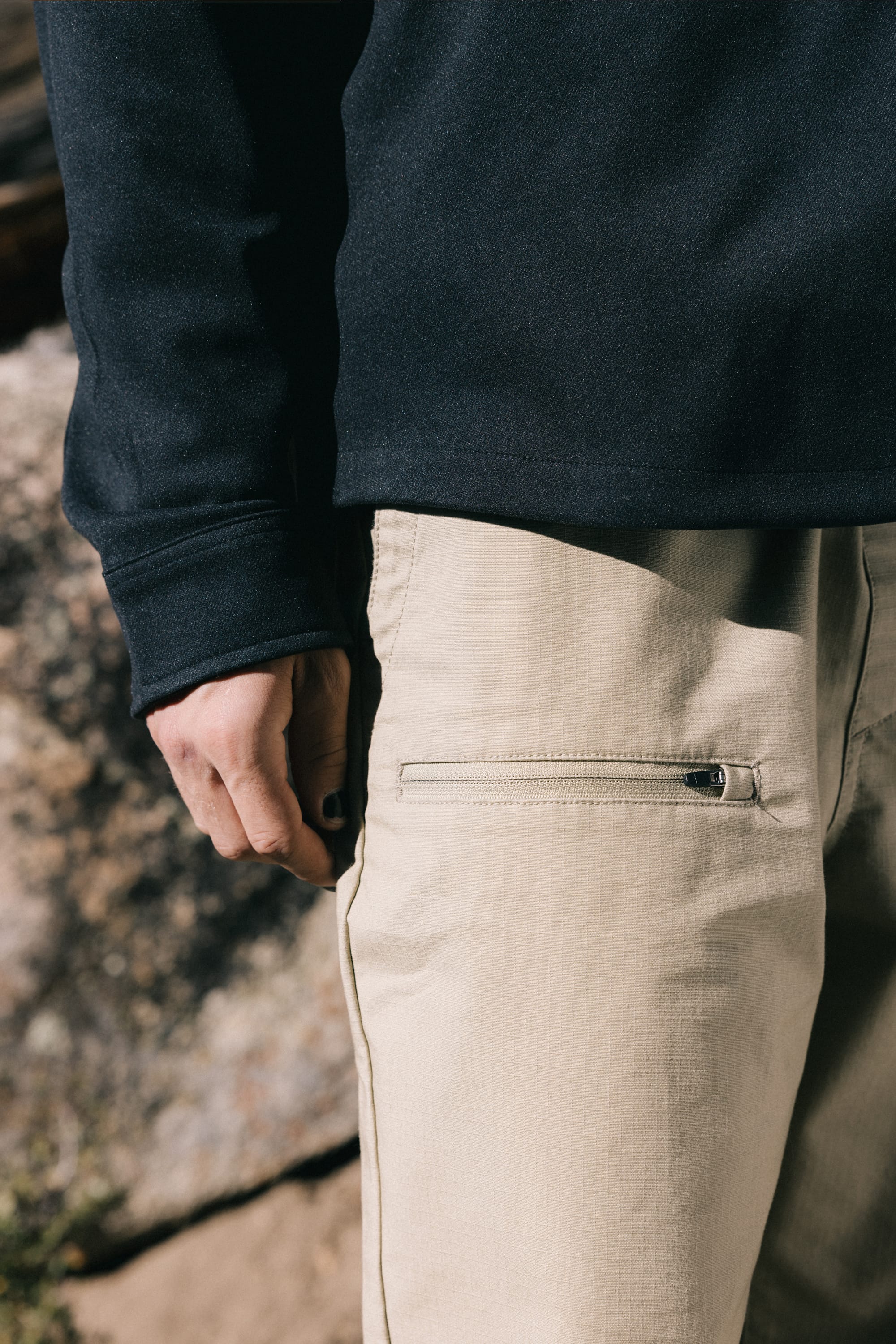 A person stands outdoors wearing a black long-sleeve shirt and the 686 Men's Free Loader Pant - Loose Fit in beige, featuring a zippered pocket. These stylish pants from 686 are designed with spandex woven fabric for enhanced comfort and flexibility, making them perfect for everyday wear.